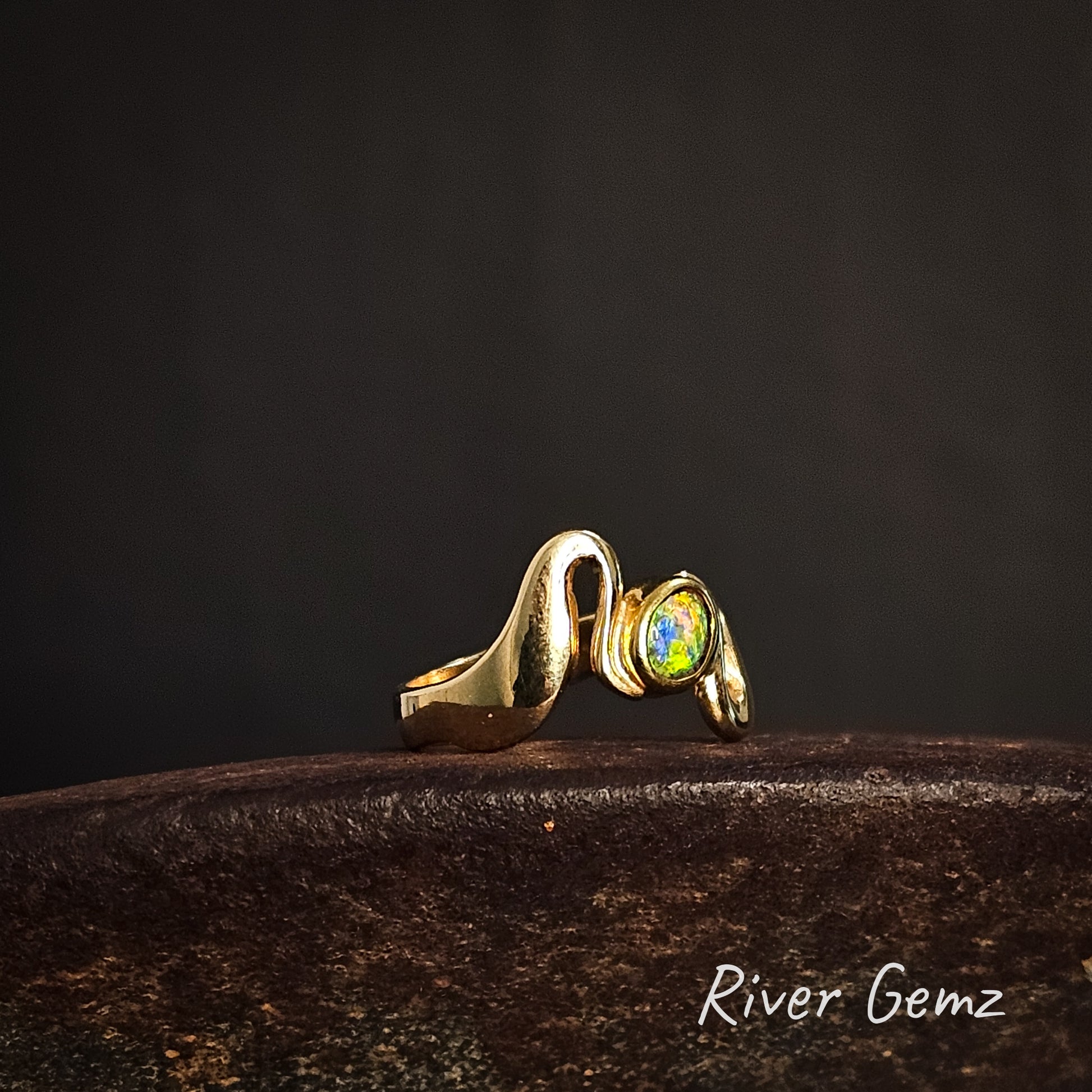 Gold ring with a colorful gemstone on a dark background, featuring the brand 'River Gemz'.