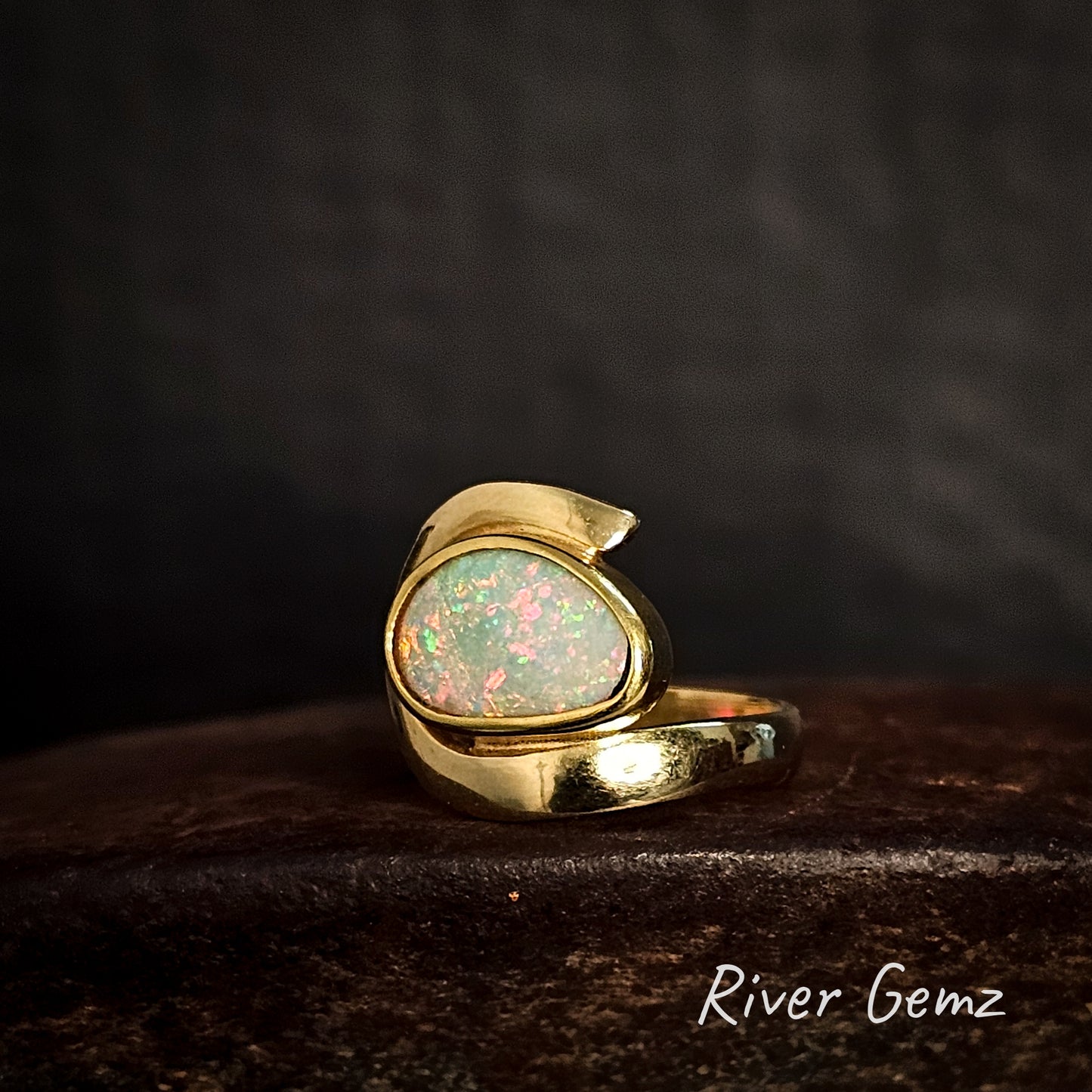 Gold ring with an opal stone on a dark background, branded 'River Gemz'.