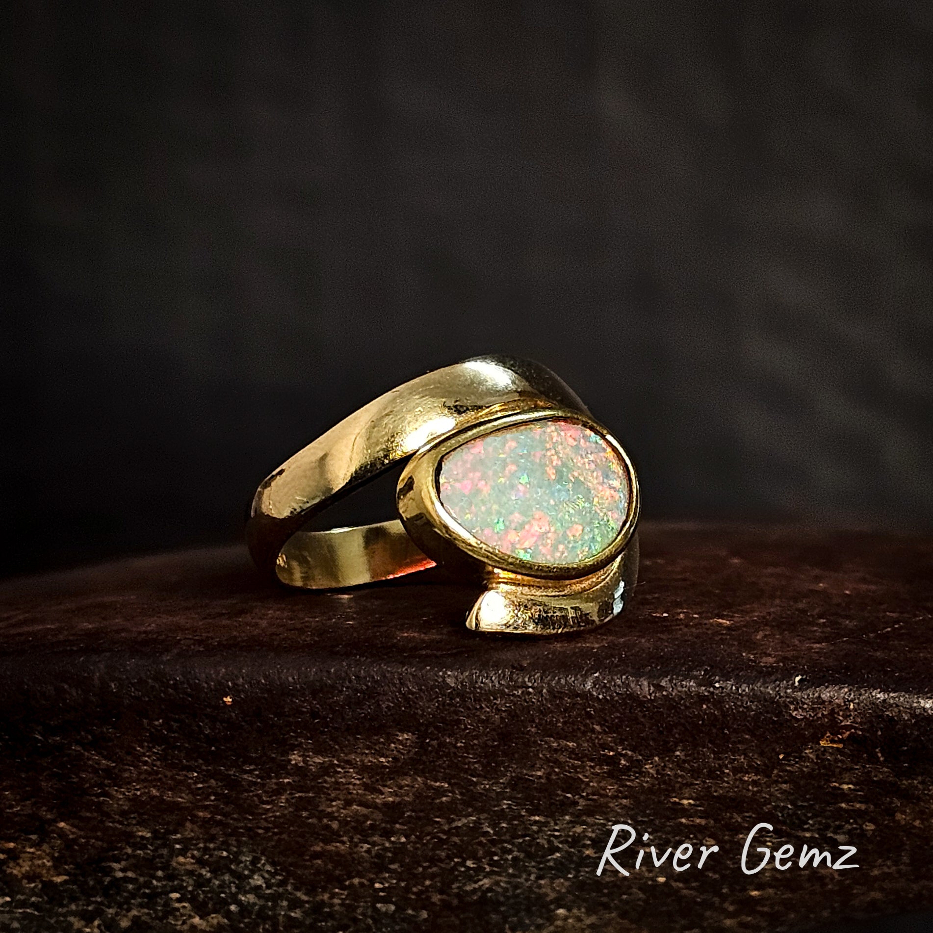 Gold ring with a multi-coloured crystal opal on a dark surface, branded 'River Gemz'.
