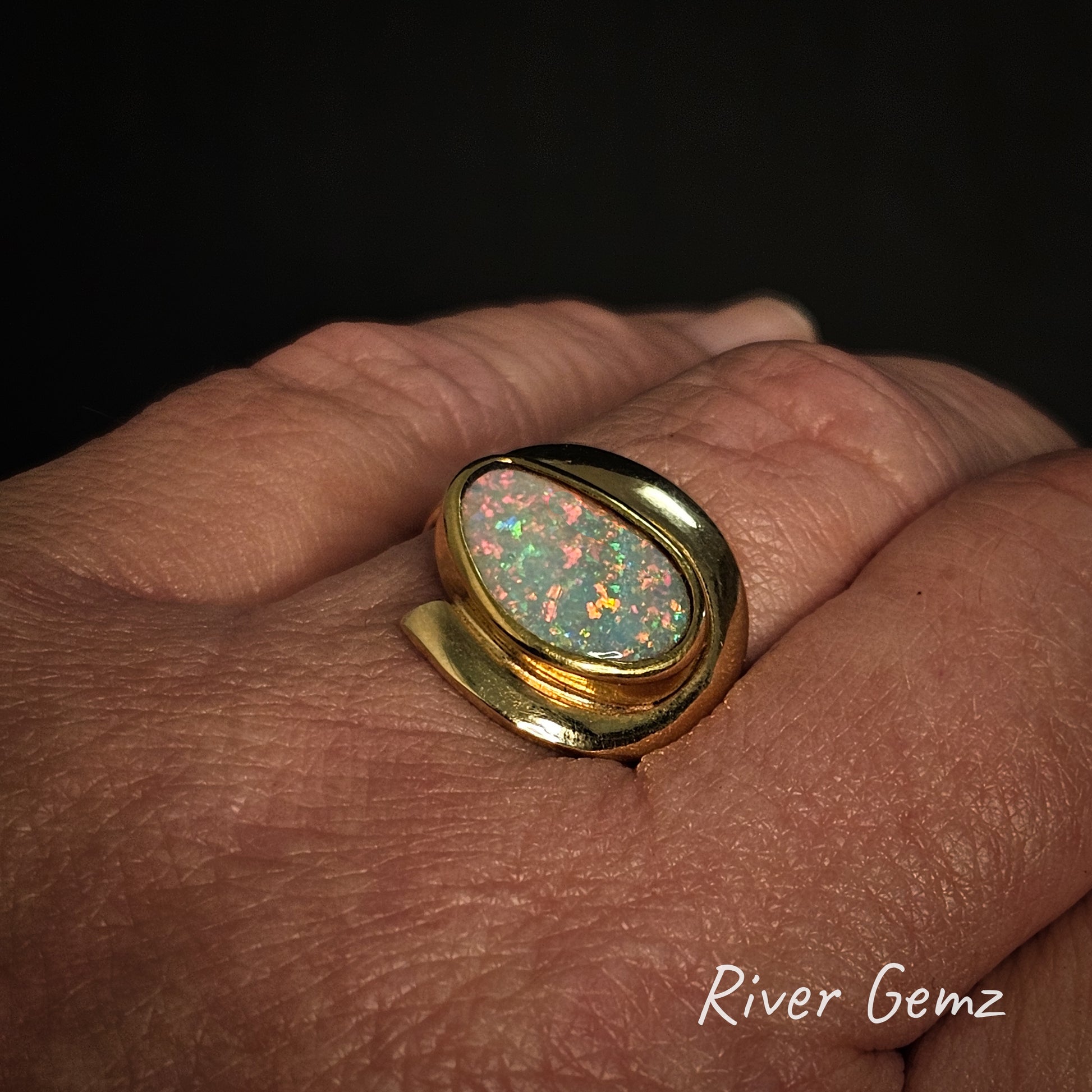 Gold ring with a colorful light opal on a hand, branded 'River Gemz'.