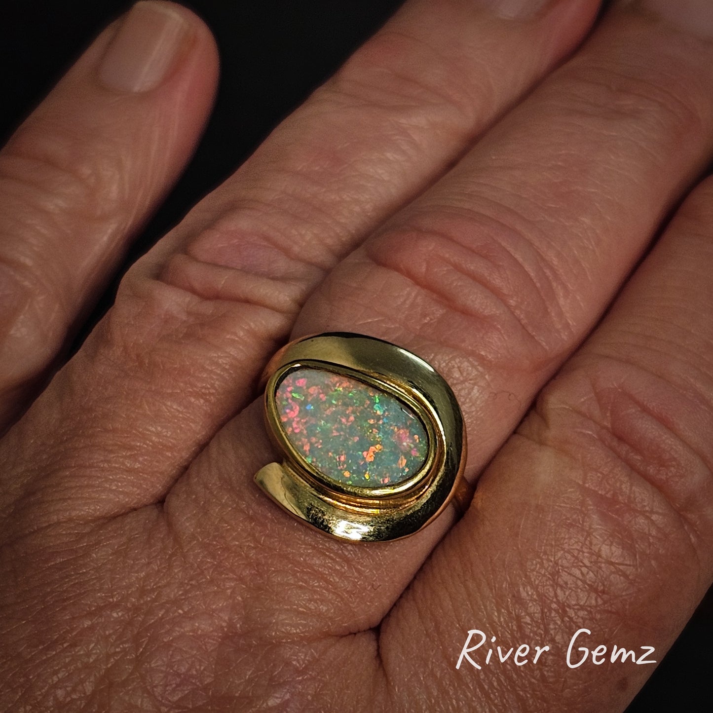 Gold ring with a colorful gemstone on a the middle finger of a hand, branded 'River Gemz'.