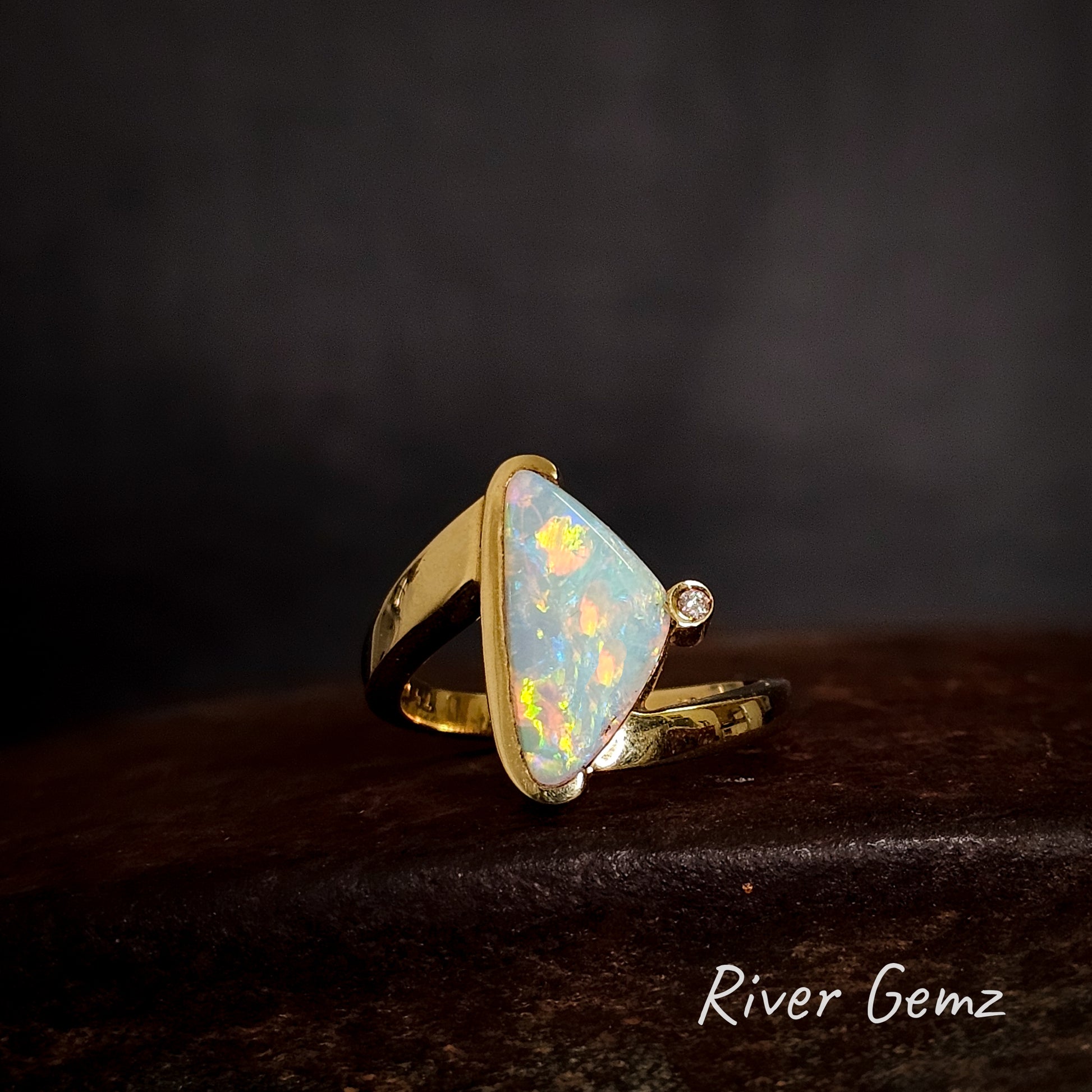 Gold ring with a large multi-coloured light opal on a dark background, branded 'River Gemz'.