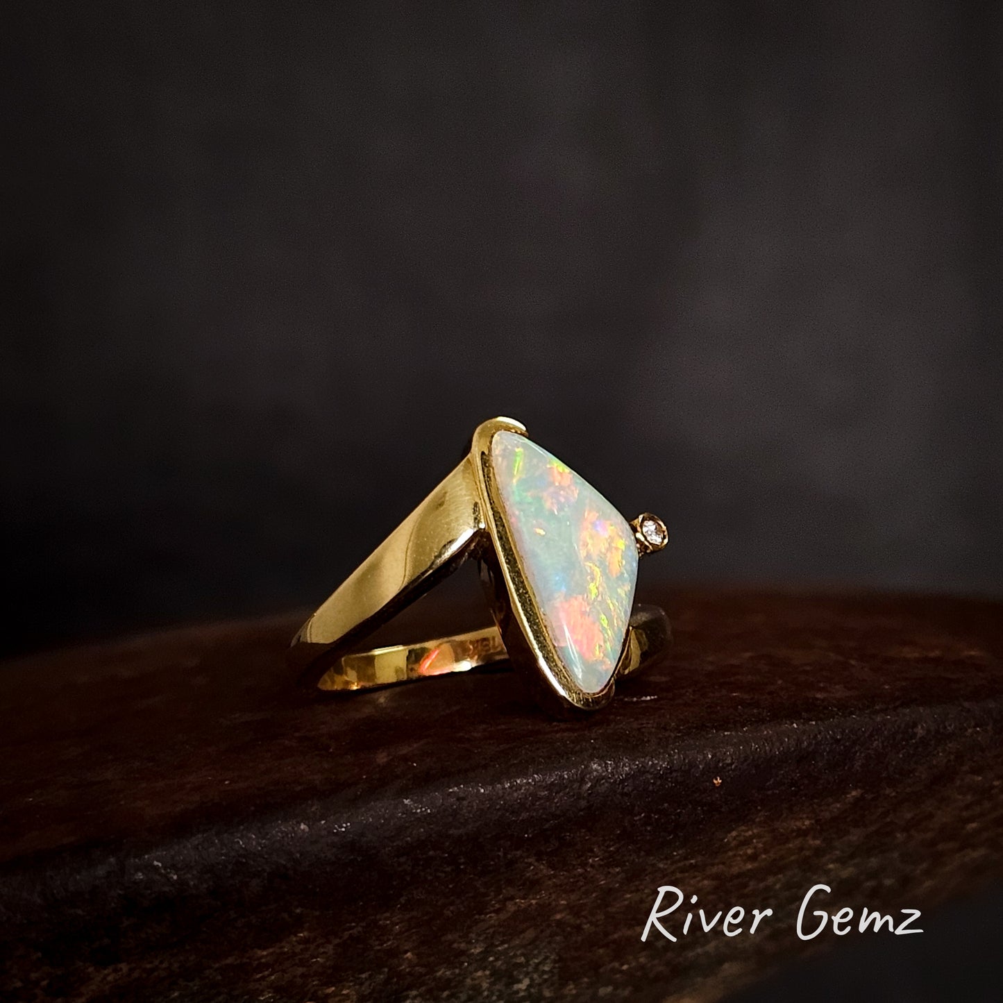 Gold ring with a triangular opal stone and small diamond on a dark background, branded 'River Gemz'.