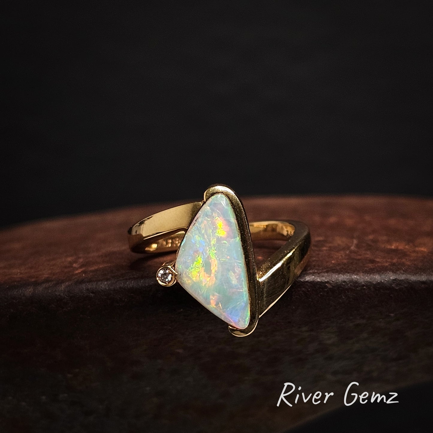 Gold ring with a triangular crystal opal stone and diamond on a dark background, branded 'River Gemz'.