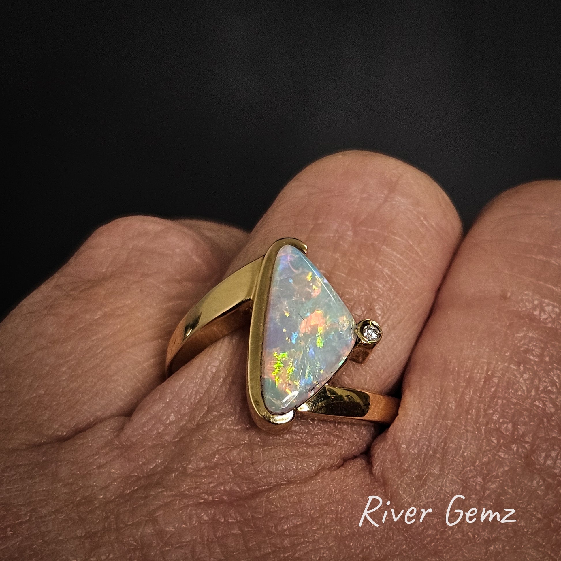 Gold ring with a colorful crystal opal on a finger, branded 'River Gemz'.