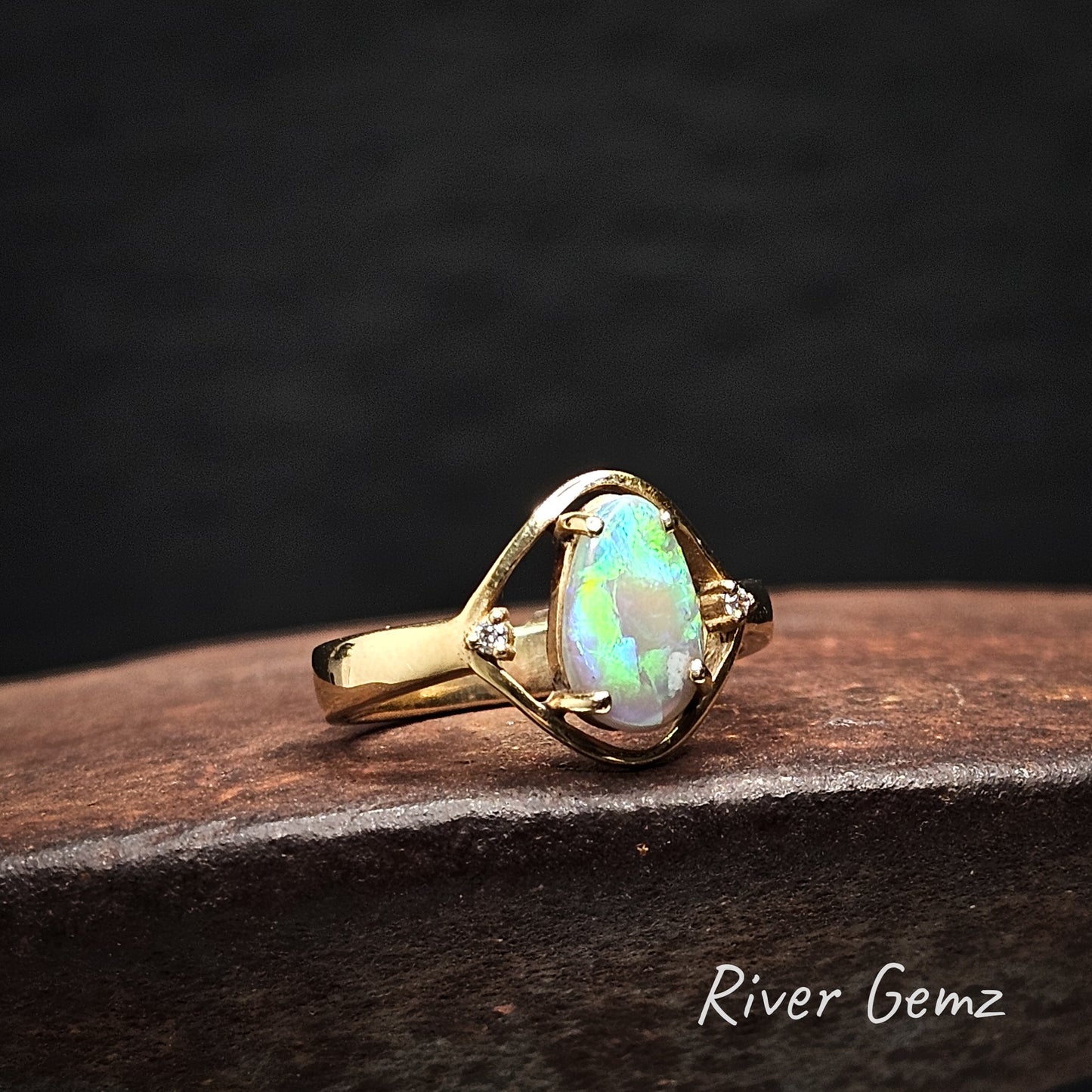 Gold ring with a tear-drop shaped light opal and a white diamond on each shoulder on a dark surface, branded 'River Gemz'.