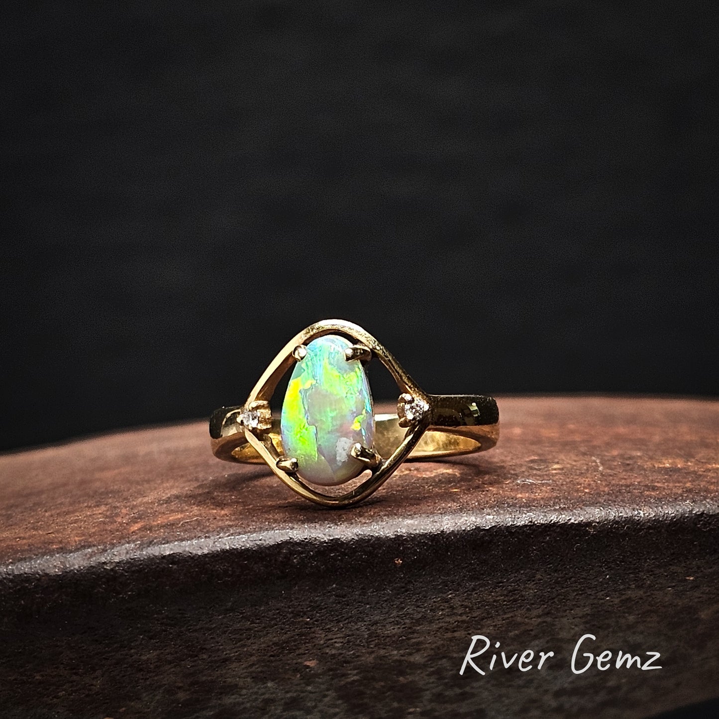 Gold ring with a colorful gemstone on a dark surface, branded 'River Gemz'.