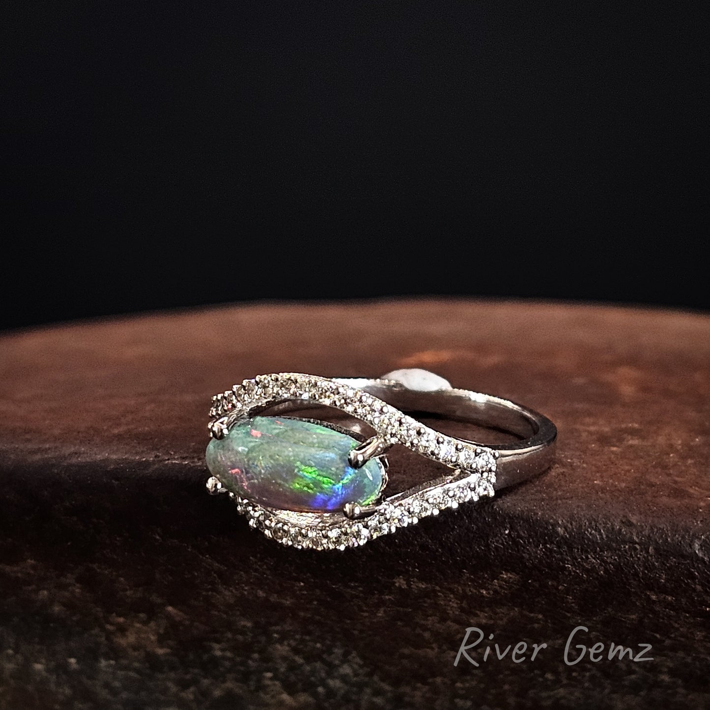 White gold ring with a claw set oval-shaped multi-coloured  opal stone surrounded by diamonds on a dark background.