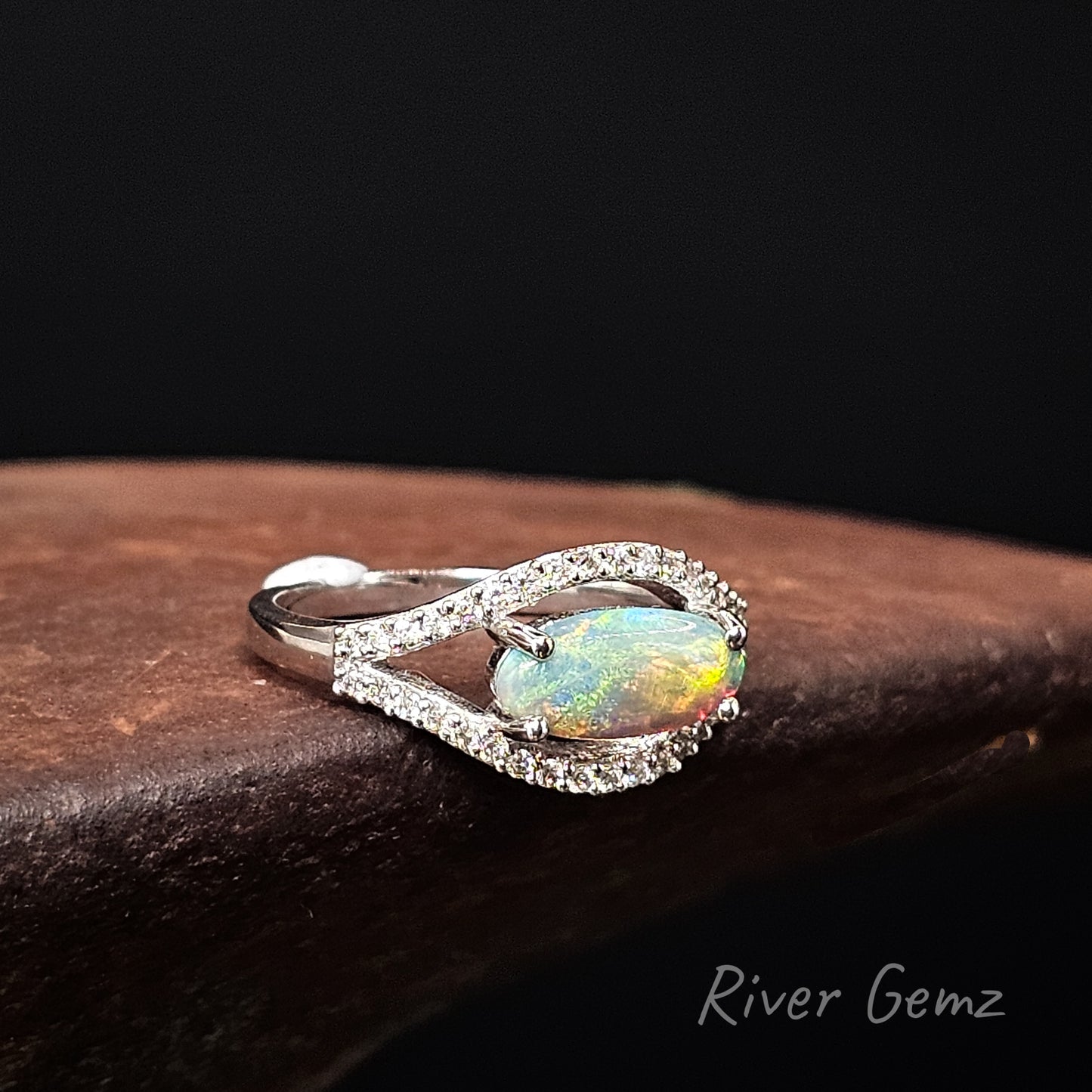 Opal ring with diamond accents on a dark surface, branded 'River Gemz'.