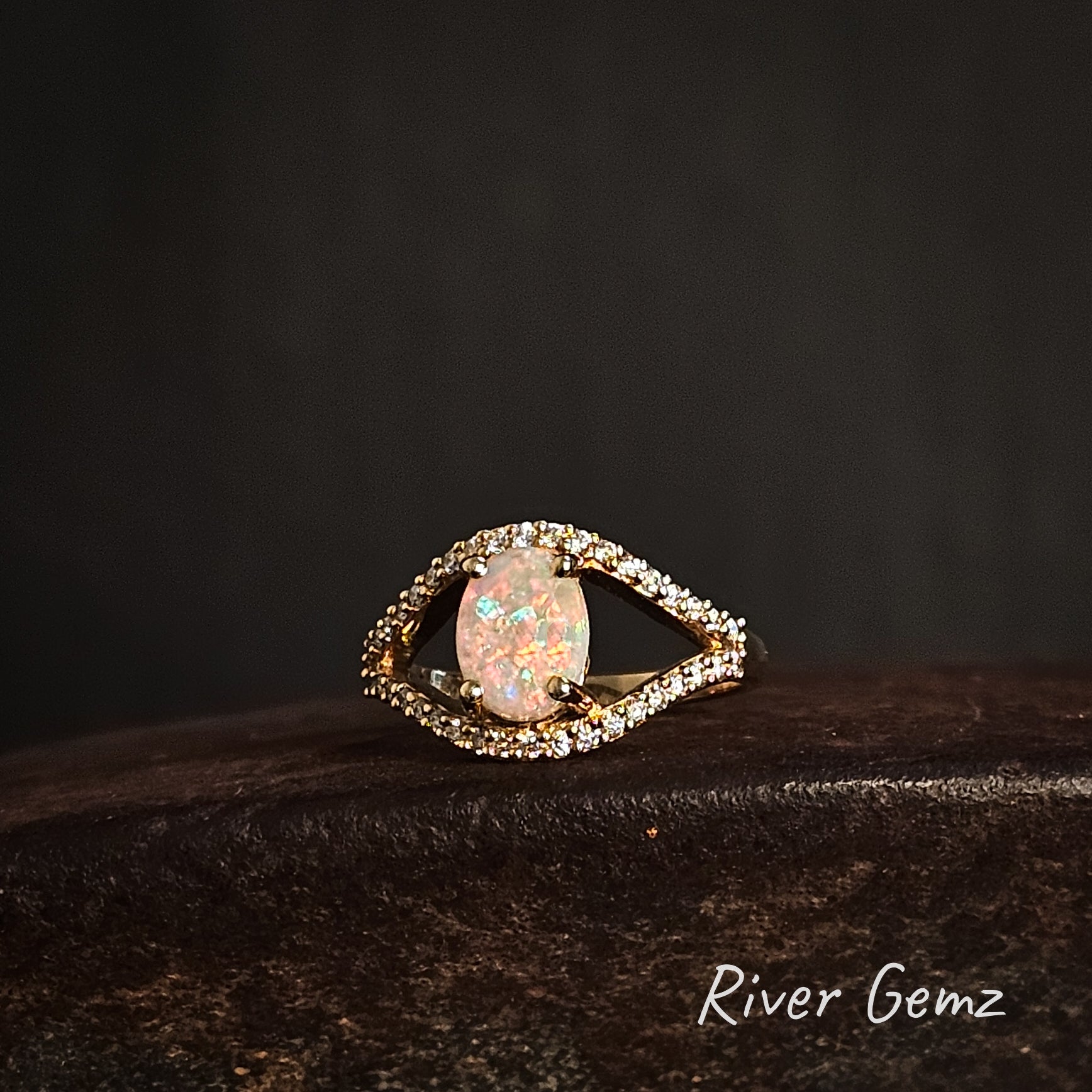Yellow gold ring in the form of an eye with a central crystal opal stone and diamond accents on a dark background, branded 'River Gemz'.
