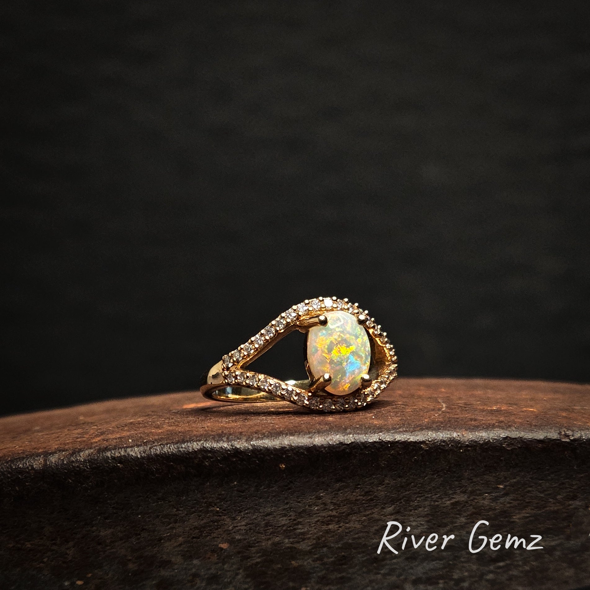 Gold ring with a multi-coloured oval shaped light opal stone and channel set diamonds on a dark background, branded 'River Gemz'.