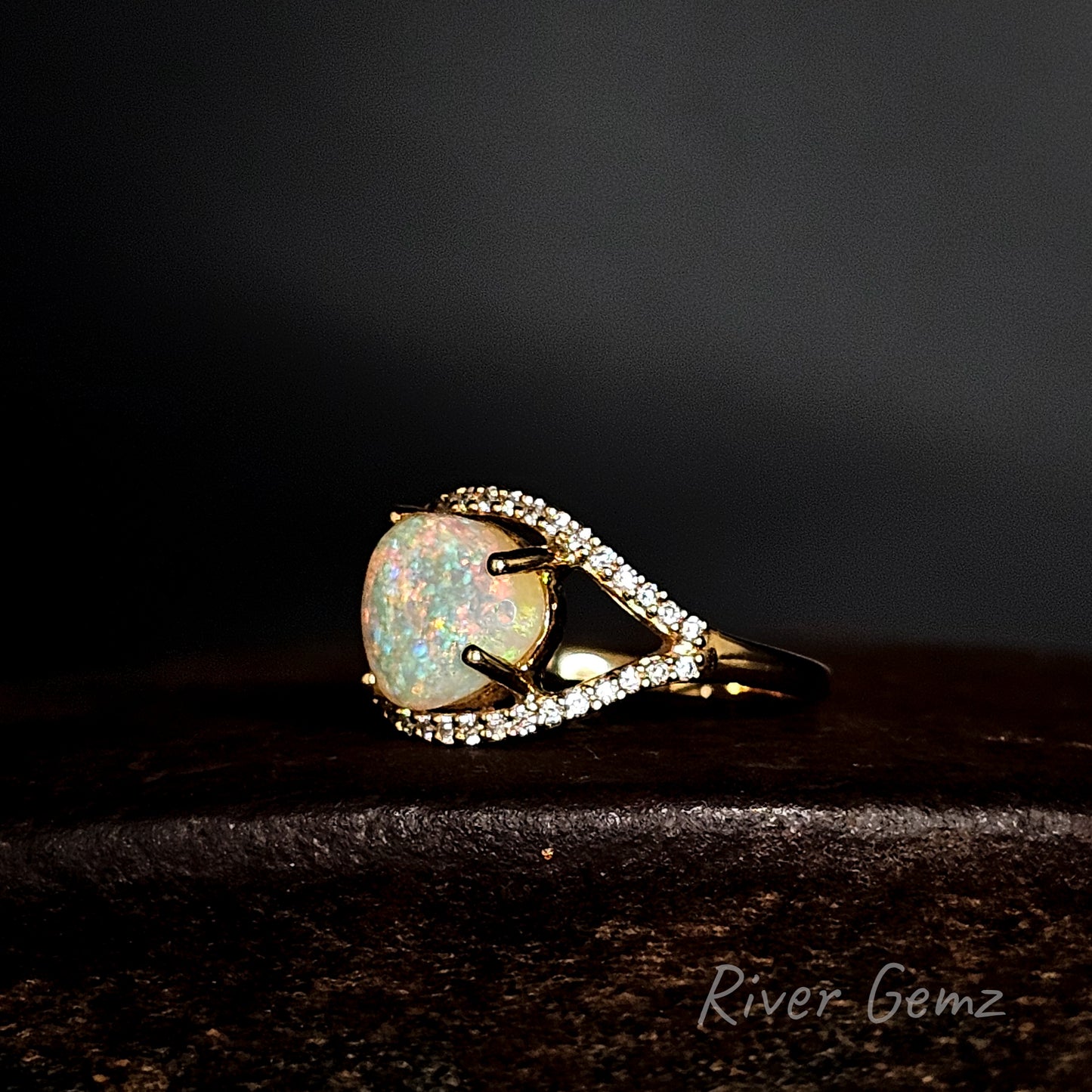 Ring with opal stone and halo of white diamonds set in gold on a dark background, featuring 'River Gemz' branding.