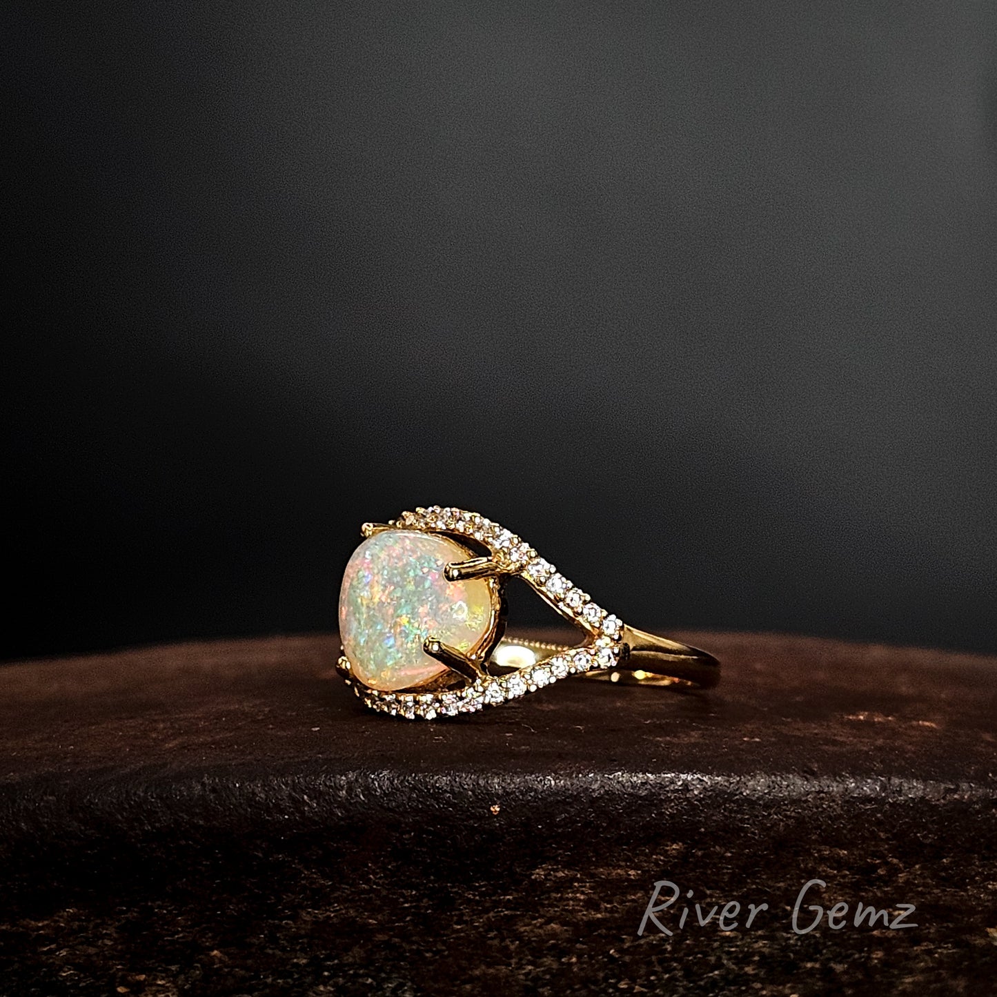 Gold ring with a large opal stone on a dark surface, branded 'River Gemz'.