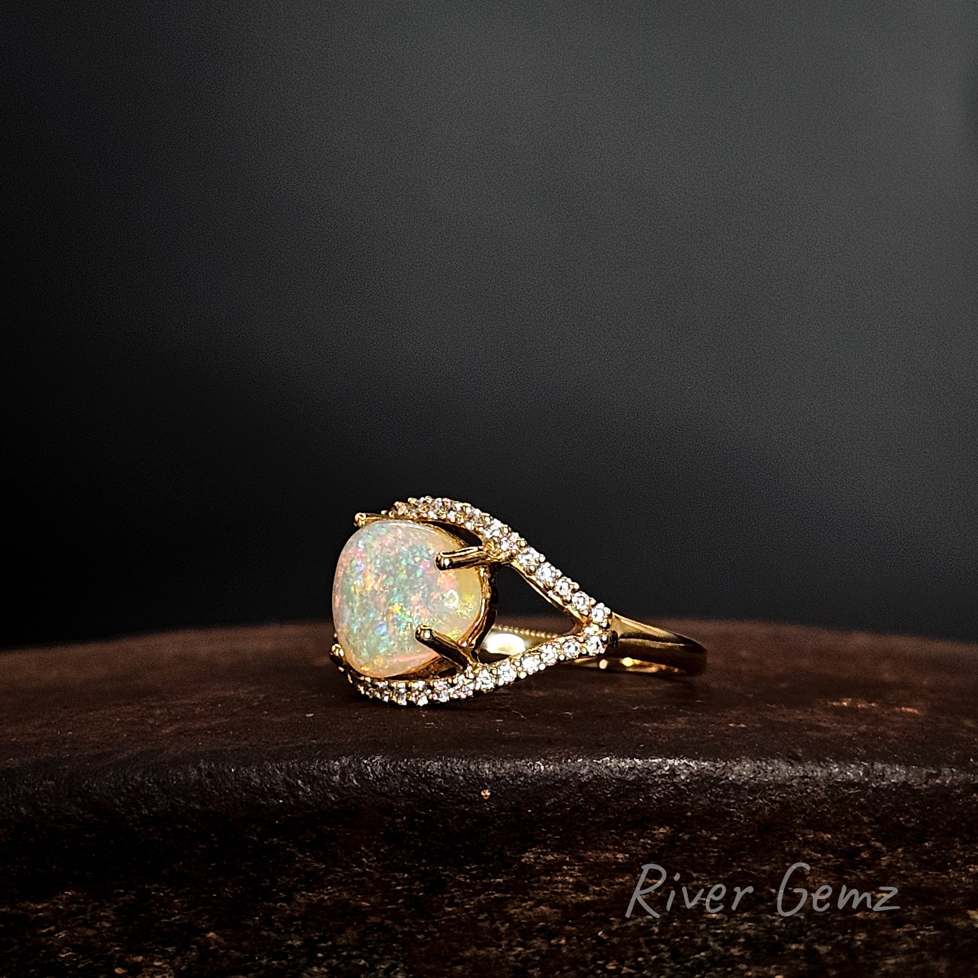 Gold ring with a large opal stone on a dark surface, branded 'River Gemz'.