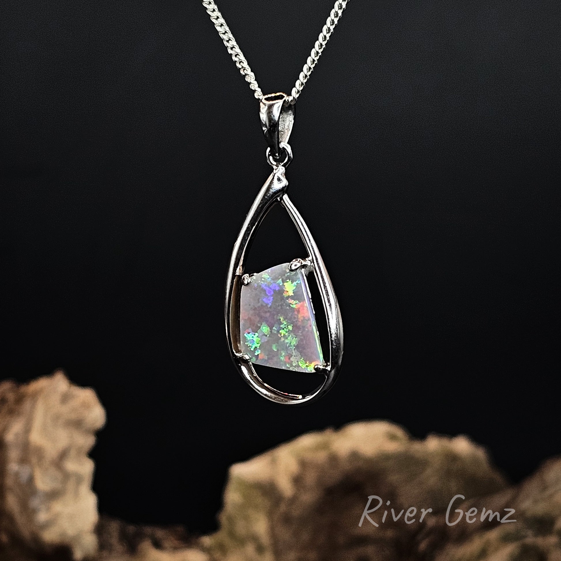 A silver teardrop pendant necklace featuring a square iridescent opal gemstone, suspended against a dark background with decorative driftwood at the bottom. The brand name 'River Gemz' is visible in the lower right corner.