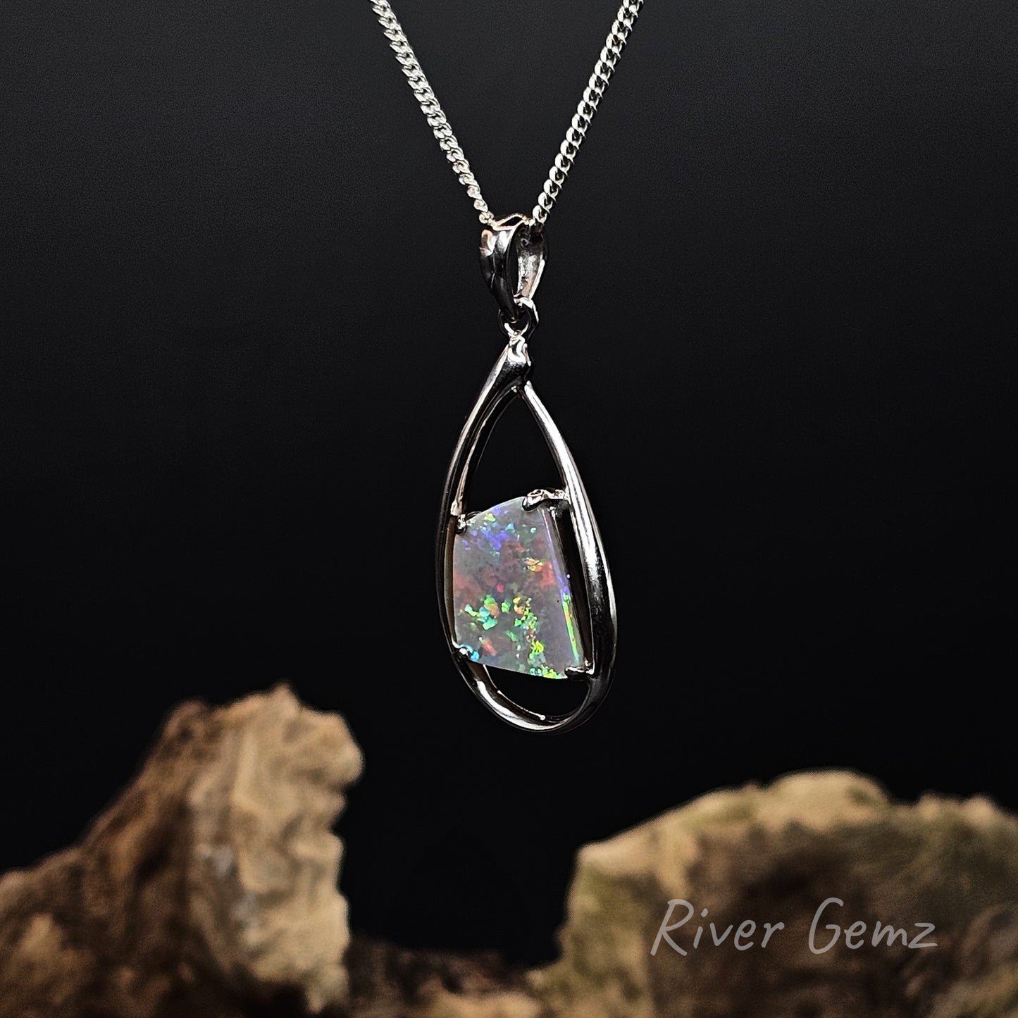 A silver teardrop pendant necklace featuring a square iridescent opal gemstone, displayed against a dark background with decorative driftwood and the 'River Gemz' logo.
