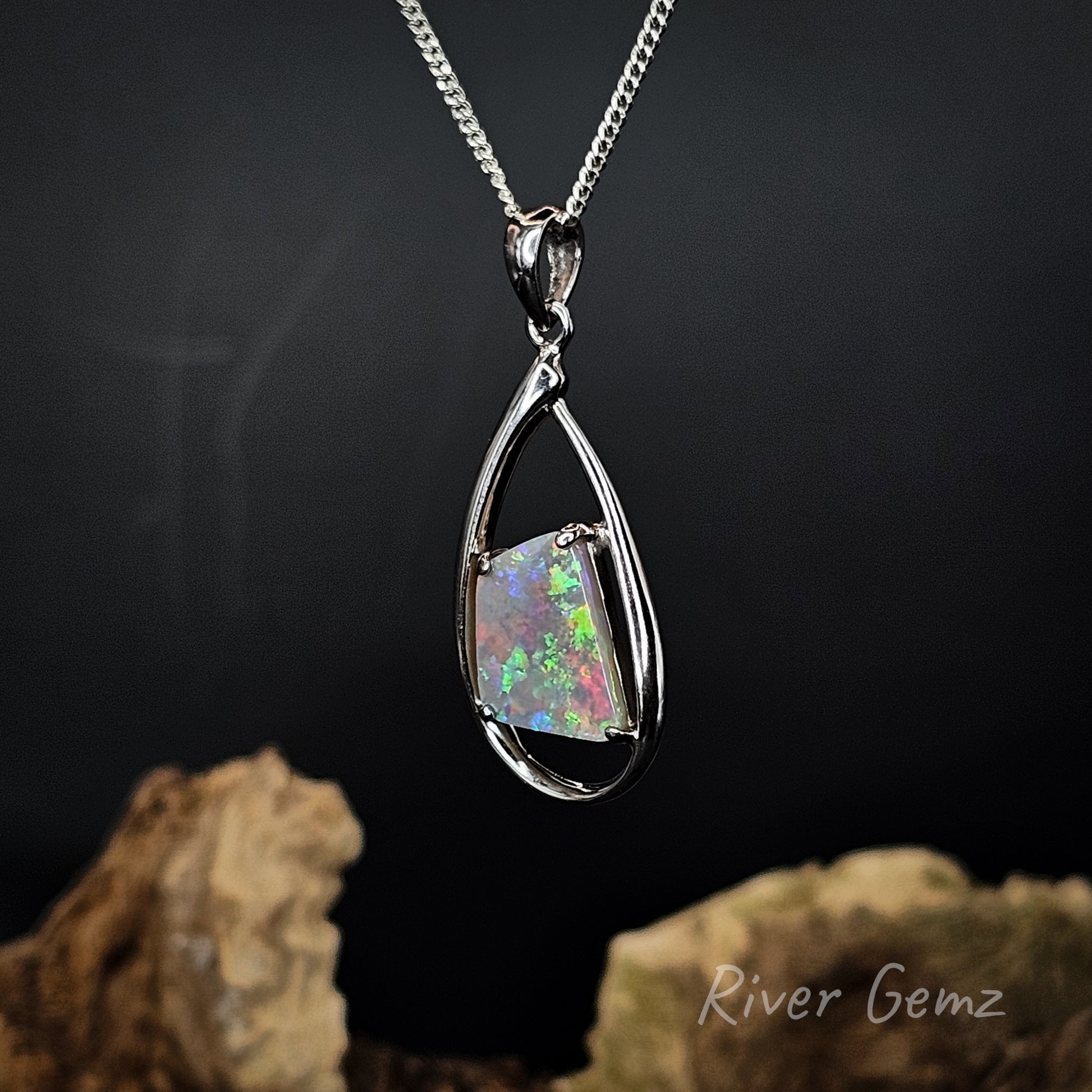 A silver teardrop pendant necklace featuring a square iridescent opal gemstone with vibrant green, pink, and blue flashes, hanging against a dark background with decorative rocks at the bottom. The brand name 'River Gemz' is visible in the bottom right corner.
