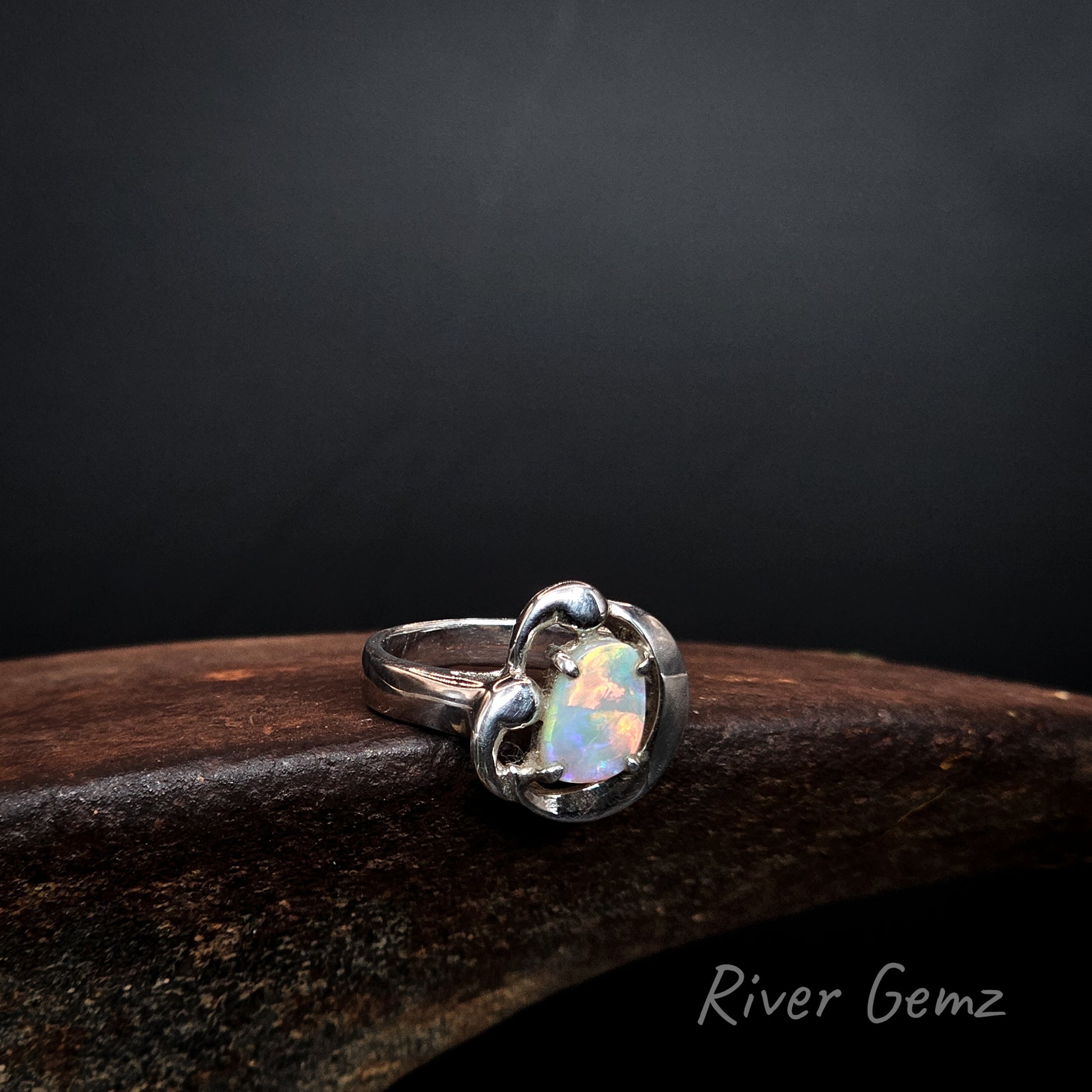 Fire colours in crystal opal claw set in silver ring on a rusty shoe anvil and dark background with 'River Gemz' branding.