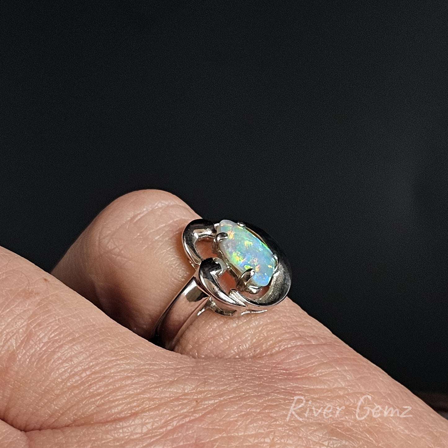A sterling silver ring featuring a round, vibrant opal gemstone, shown worn on a person's finger against a dark background. The brand name 'River Gemz' is watermarked in the bottom right corner.