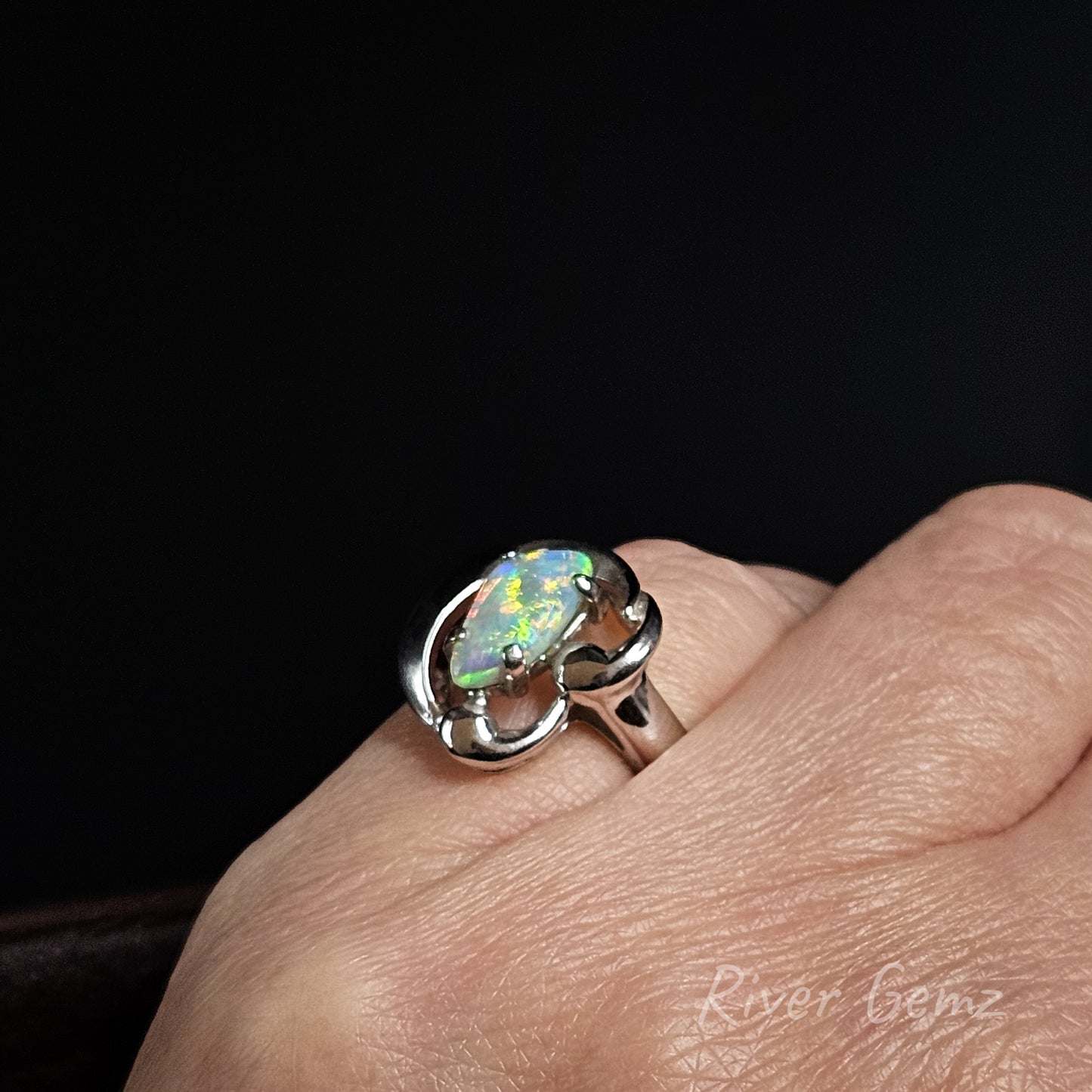 A silver ring featuring a large, vibrant iridescent opal gemstone, shown worn on a person's finger against a solid black background.