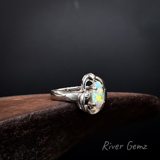 A sterling silver ring featuring an iridescent opal gemstone, resting on a dark wooden surface against a dark background. The brand name 'River Gemz' is visible in the bottom right corner.