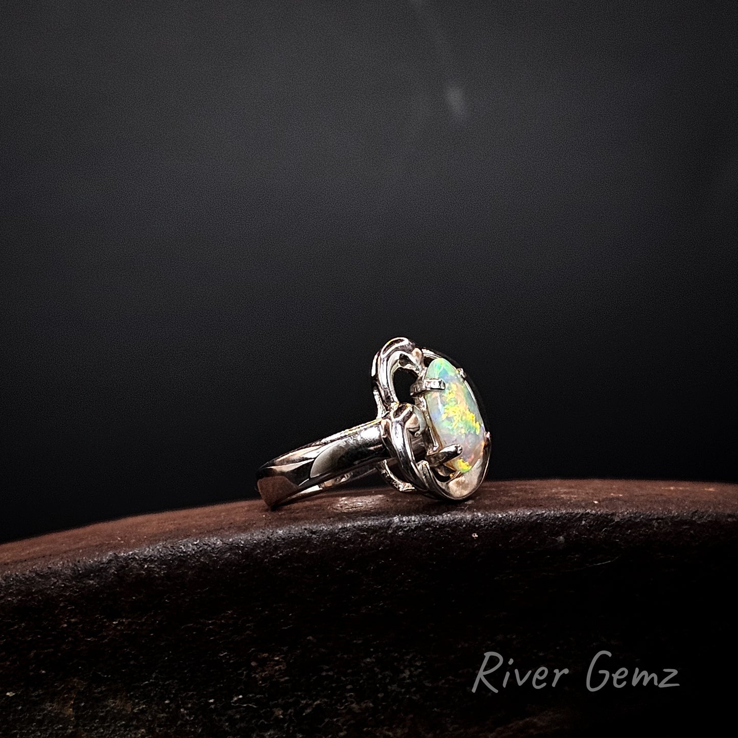 A silver ring featuring a large, round, iridescent opal gemstone, resting on a dark wooden surface against a dark background. The brand name 'River Gemz' is visible in the bottom right corner.