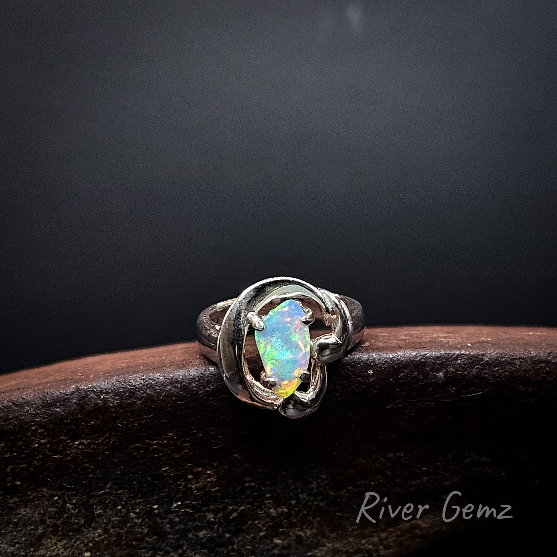 A sterling silver ring featuring a free form cut crystal opal, resting on a dark brown textured surface against a dark gray background. The brand name 'River Gemz' is visible in the bottom right corner.
