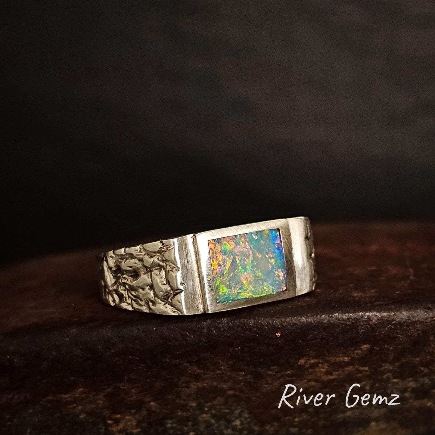 Silver ring with a rectangular shaped multi-coloured opal on a dark background, featuring 'River Gemz' branding.
