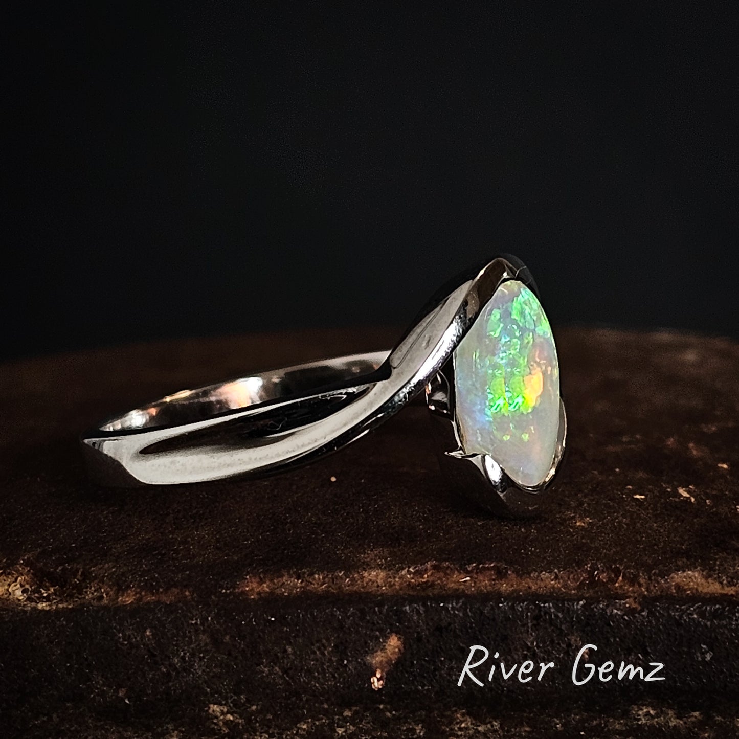 Silver ring with a partial besel set colorful opal stone on a dark background, branded 'River Gemz'.