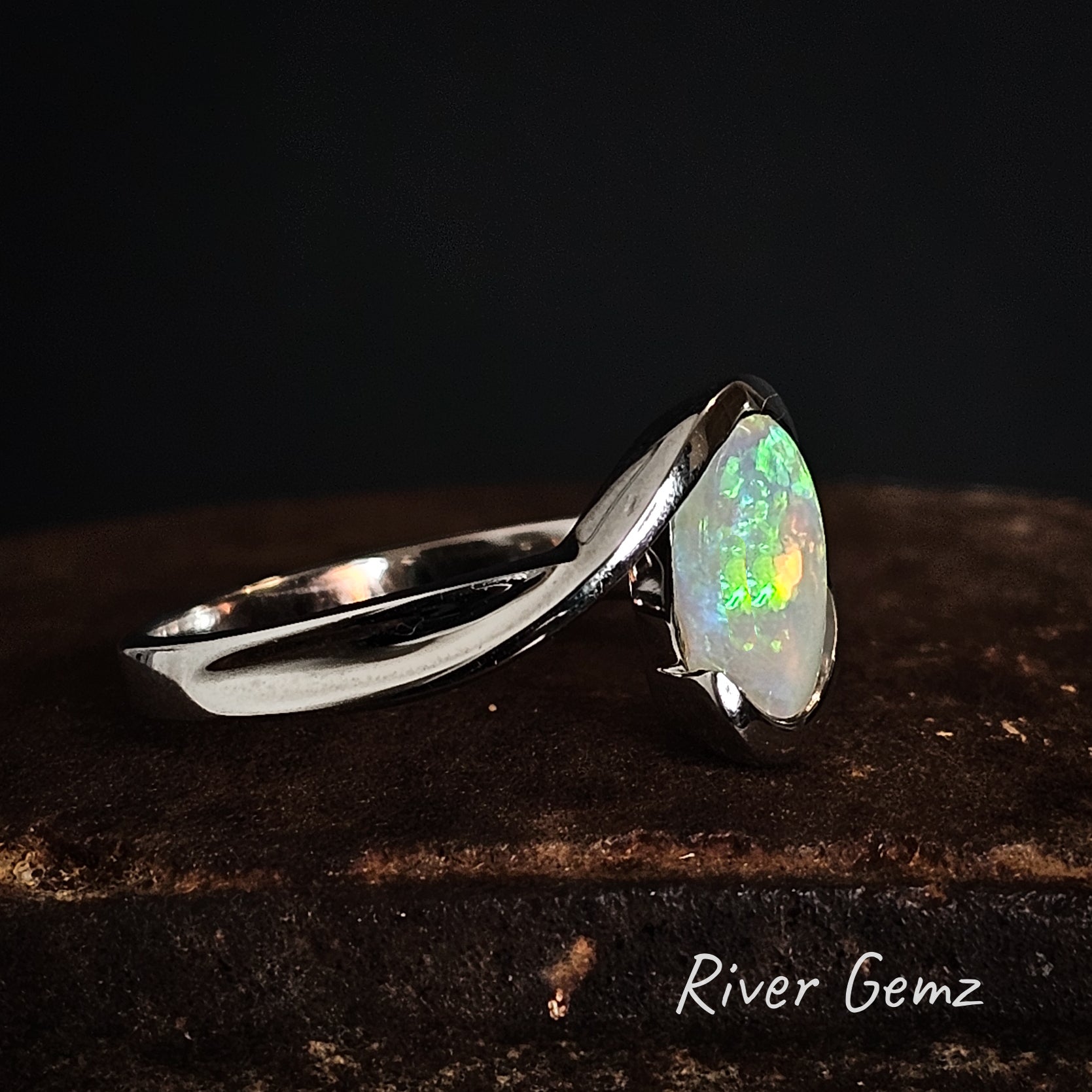 Silver ring with a partial besel set colorful opal stone on a dark background, branded 'River Gemz'.