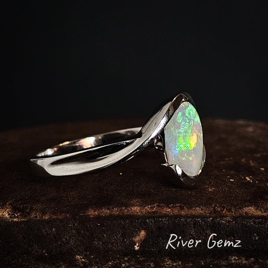 Silver ring with a partial besel set colorful opal stone on a dark background, branded 'River Gemz'.
