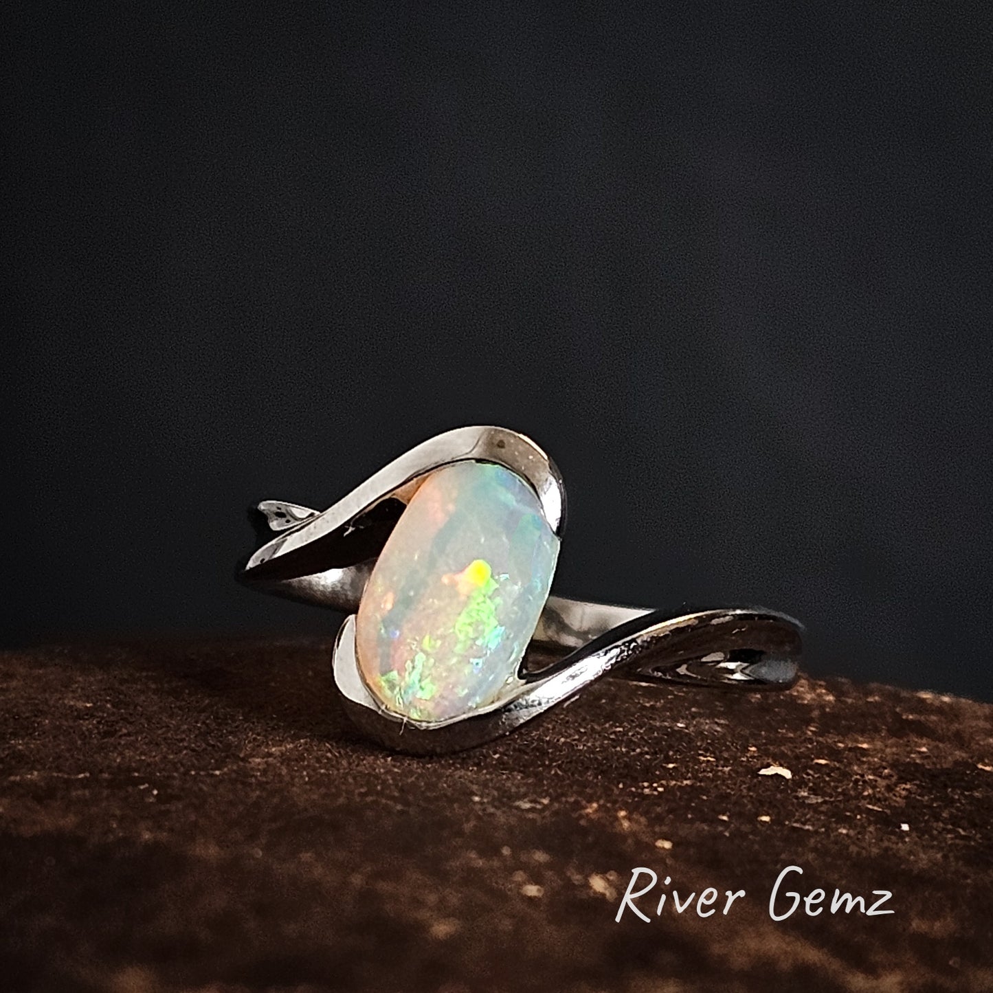 Silver ring with an oval shaped crystal opal stone on a dark background, branded 'River Gemz'.