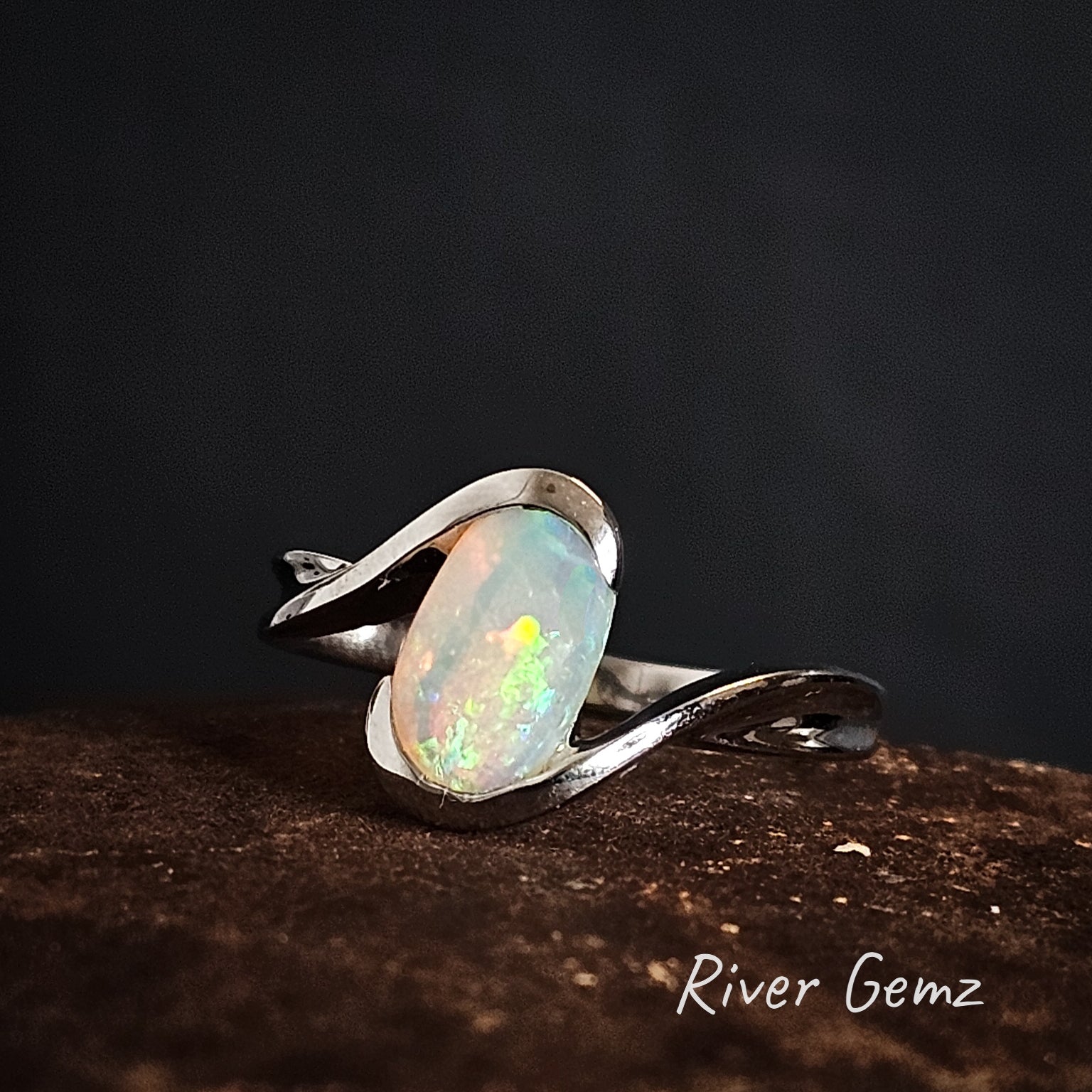 Silver ring with an oval shaped crystal opal stone on a dark background, branded 'River Gemz'.