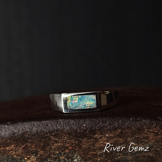 Silver ring with a greenish-blue gemstone on a dark surface, branded 'River Gemz'.