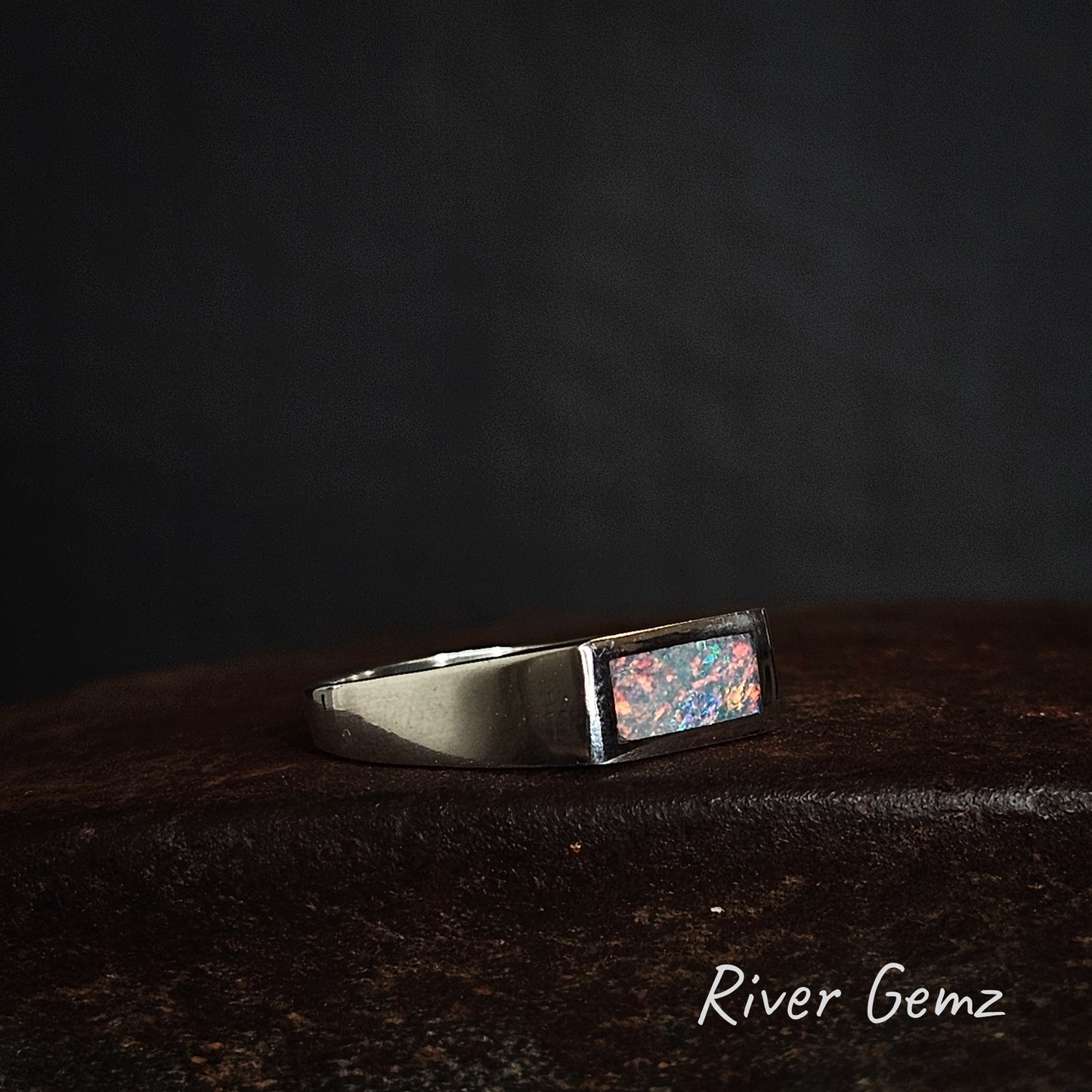 Silver ring with a rectangular shaped colourful crystal opal on a dark background, branded 'River Gemz'.