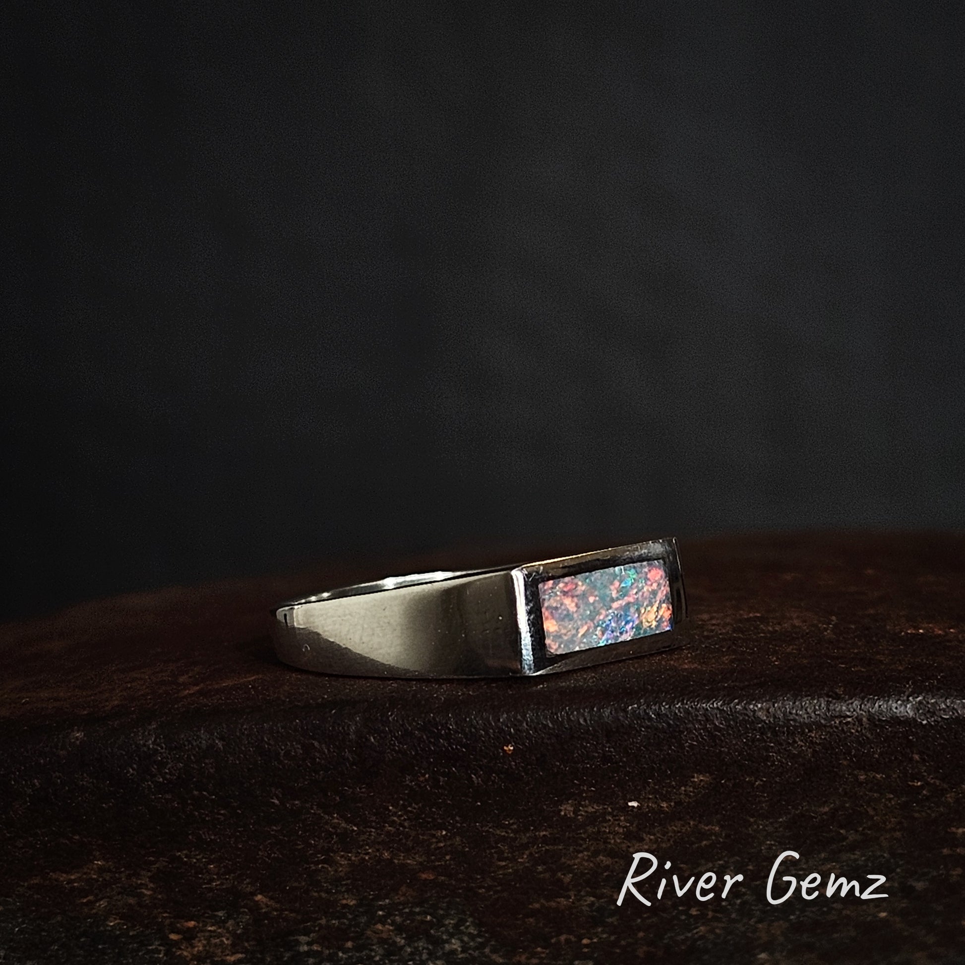 Silver ring with a rectangular shaped colourful crystal opal on a dark background, branded 'River Gemz'.