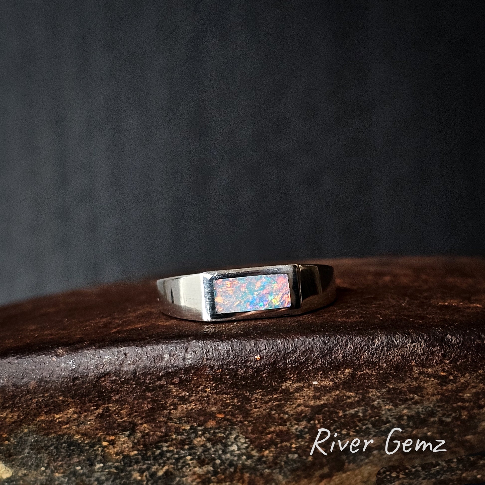 Silver ring with multicoloured opal on a dark surface, branded 'River Gemz'.