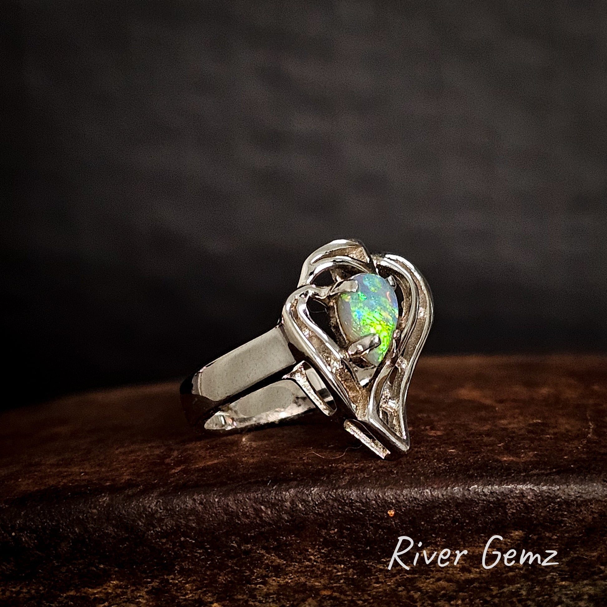 Silver ring with a heart-shaped opal stone on a dark background, branded 'River Gemz'.
