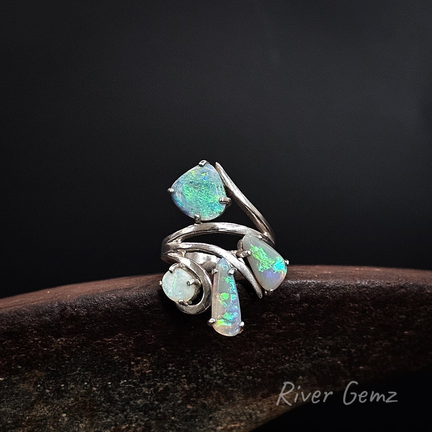 A silver ring featuring four iridescent opal gemstones in various shapes, displayed on a dark wooden surface against a black background. The brand name 'River Gemz' is visible in the bottom right corner.