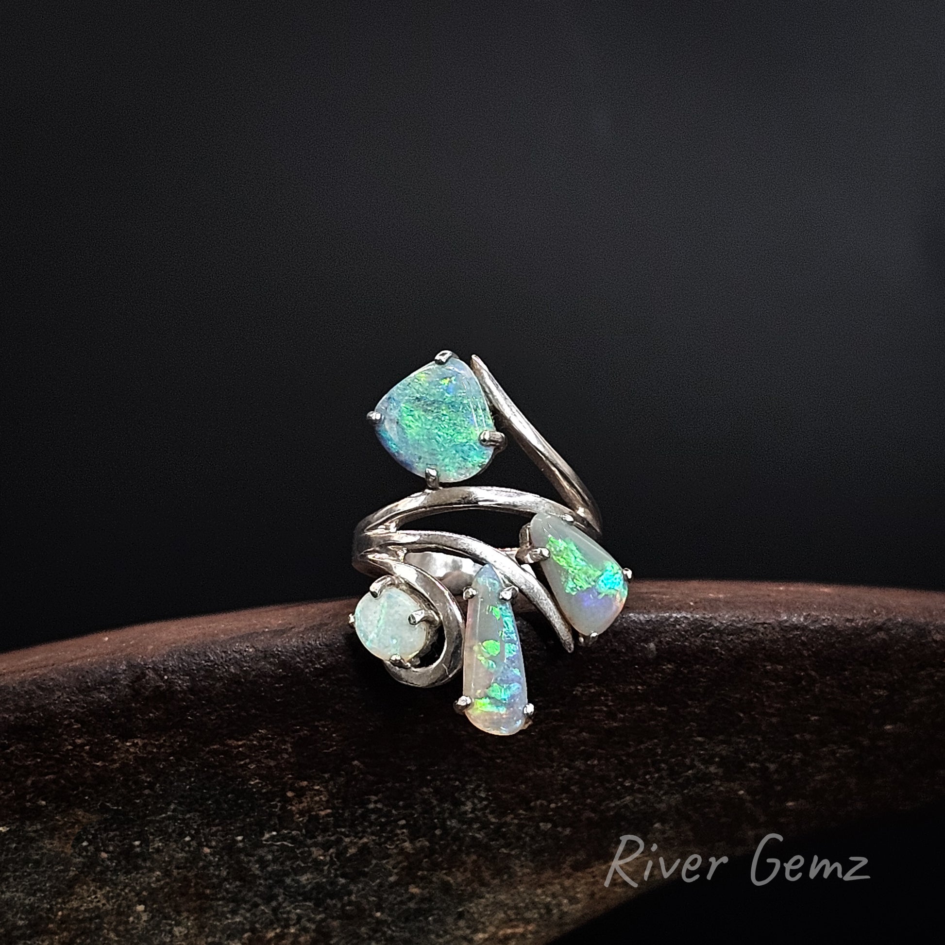 A silver ring featuring four iridescent opal gemstones in various shapes, displayed on a dark wooden surface against a black background. The brand name 'River Gemz' is visible in the bottom right corner.