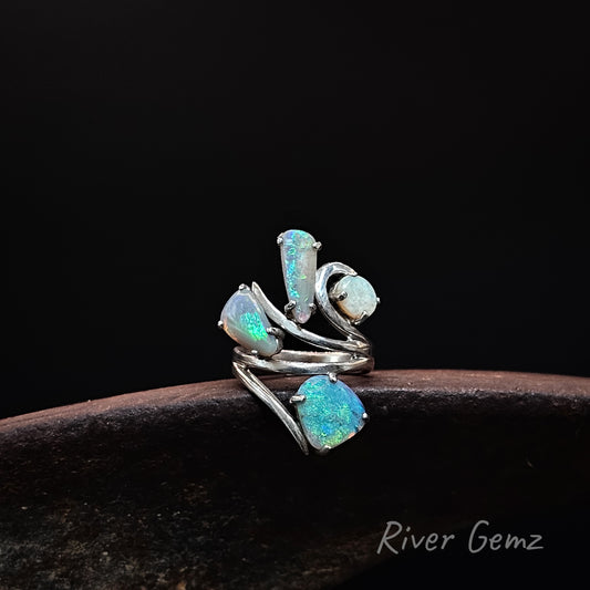 A silver wrap ring featuring four iridescent opal gemstones in different shapes, displayed on a dark wooden surface against a black background. The brand name 'River Gemz' is visible in the bottom right corner.