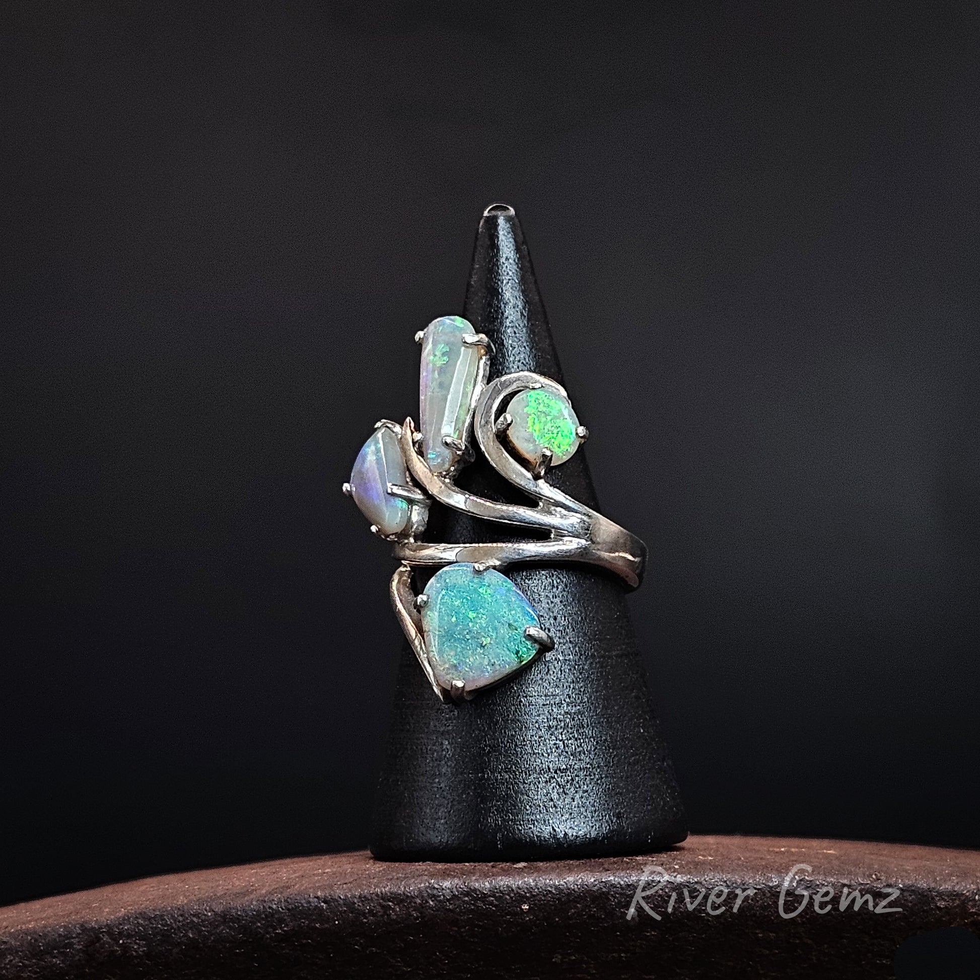 A silver ring with four teardrop-shaped opal gemstones in various shades of green, blue, and iridescent white, displayed on a black cone stand against a dark background.