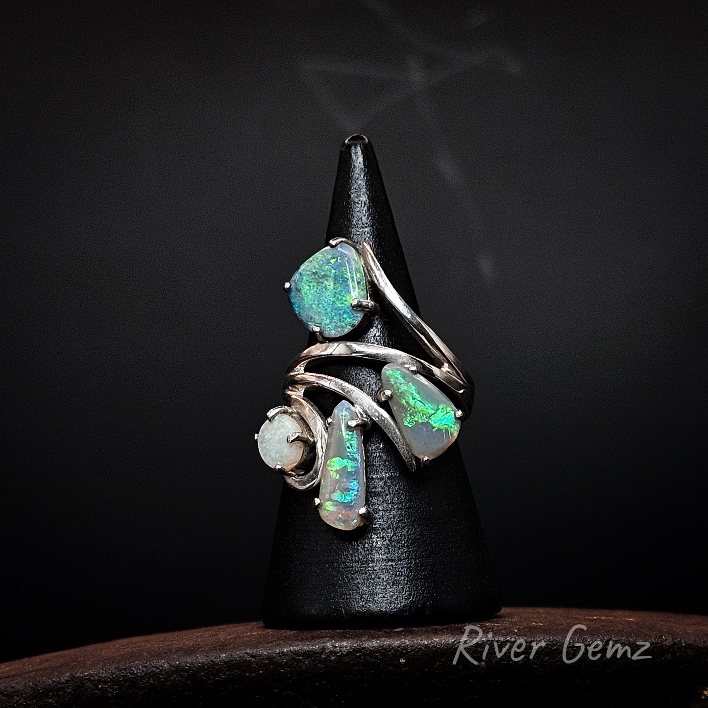 A silver ring with four opal gemstones displaying vibrant green and blue flashes, displayed on a black cone stand against a dark background.