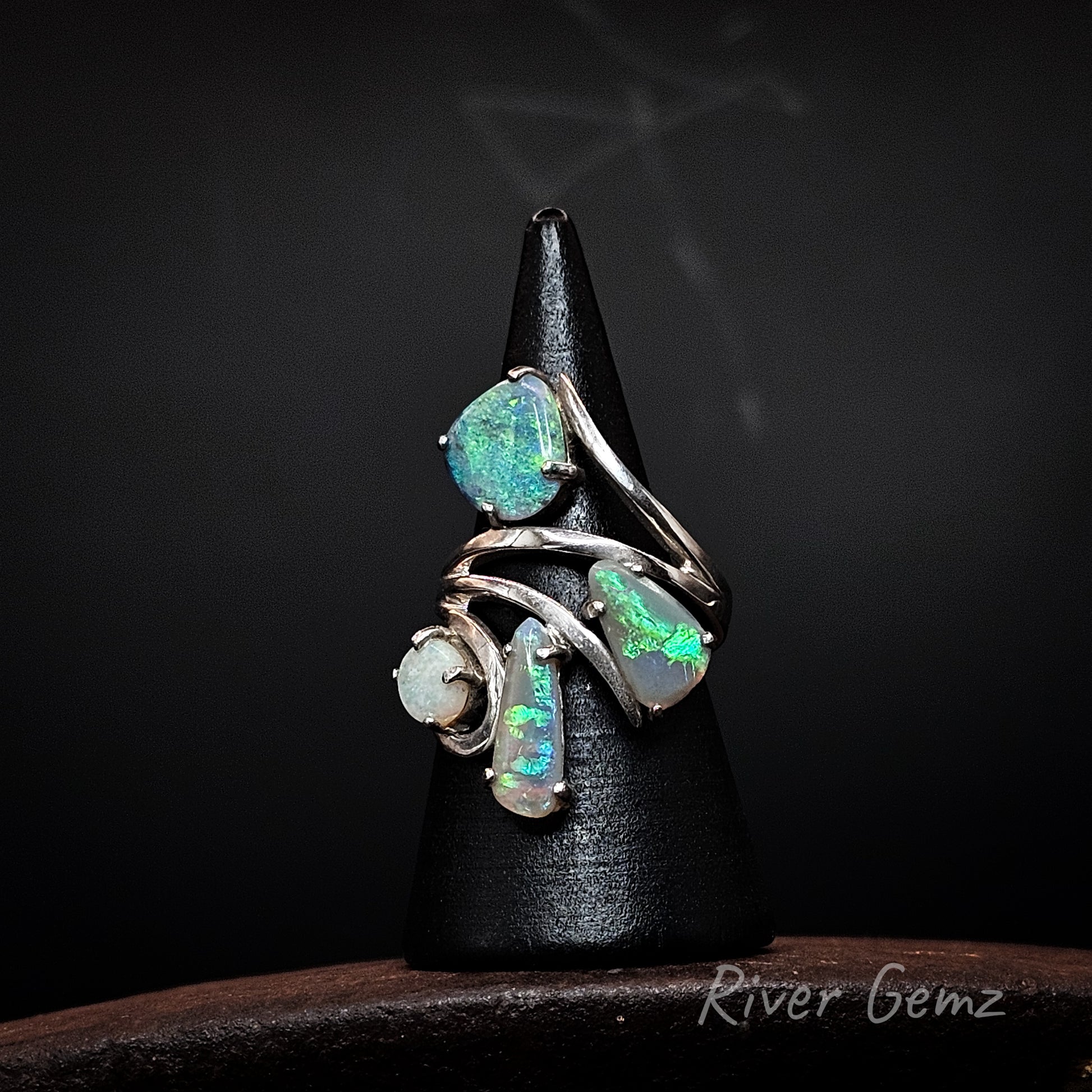 A silver ring with four opal gemstones displaying vibrant green and blue flashes, displayed on a black cone stand against a dark background.