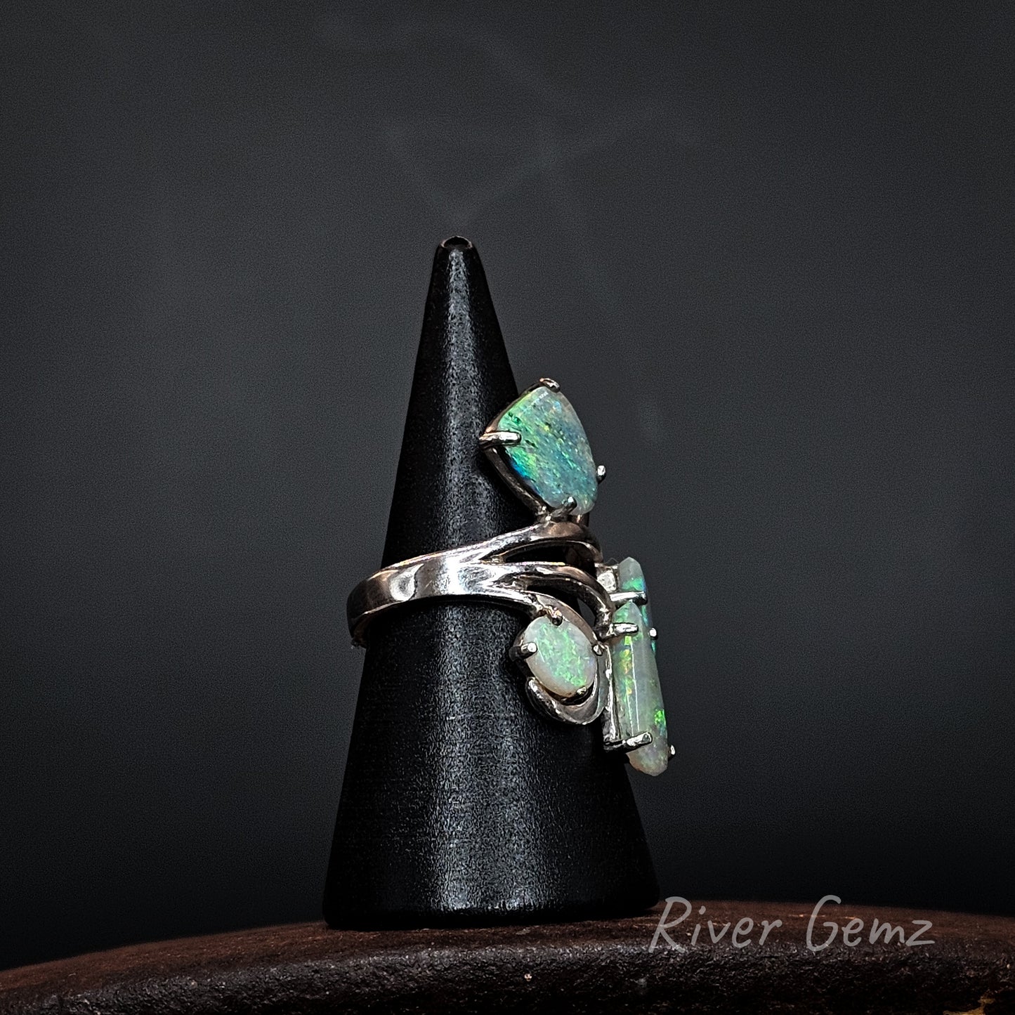 A silver ring showing three green opal stones displayed on a black cone stand against a dark background. The brand name 'River Gemz' is visible as a watermark in the bottom right corner.