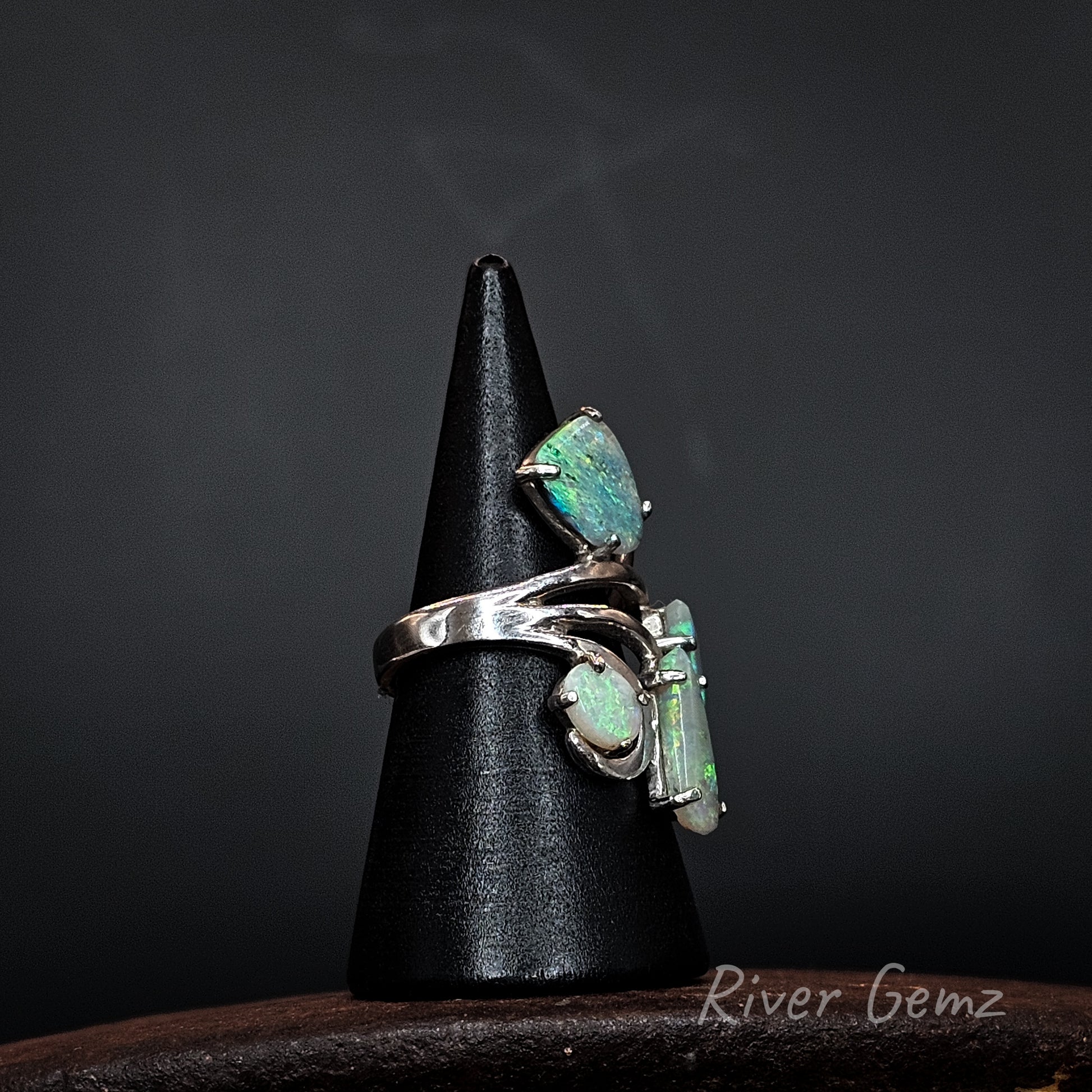 A silver ring showing three green opal stones displayed on a black cone stand against a dark background. The brand name 'River Gemz' is visible as a watermark in the bottom right corner.