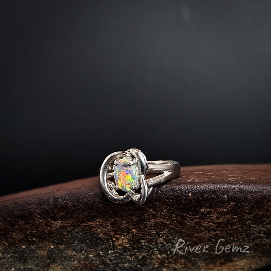 A sterling silver ring with a colorful, iridescent opal gemstone, resting on a dark brown textured surface against a dark gray background. The brand name 'River Gemz' is visible as a watermark in the bottom right corner.