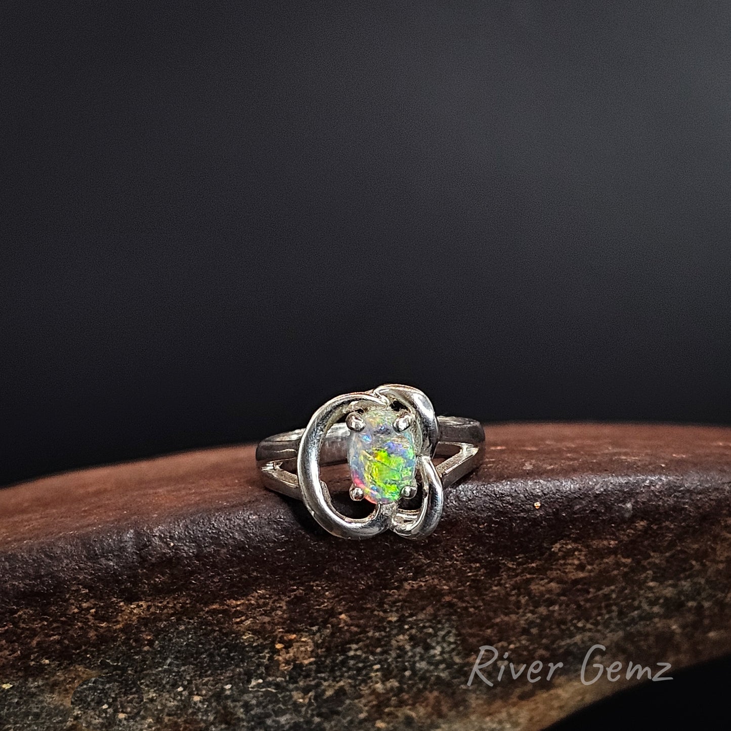 A silver ring featuring a round iridescent opal gemstone in a prong setting, surrounded by a polished silver band with two interlocking curved loops. The ring is displayed on a dark brown wooden surface against a dark gray background, with a 'River Gemz' watermark in the bottom right corner.