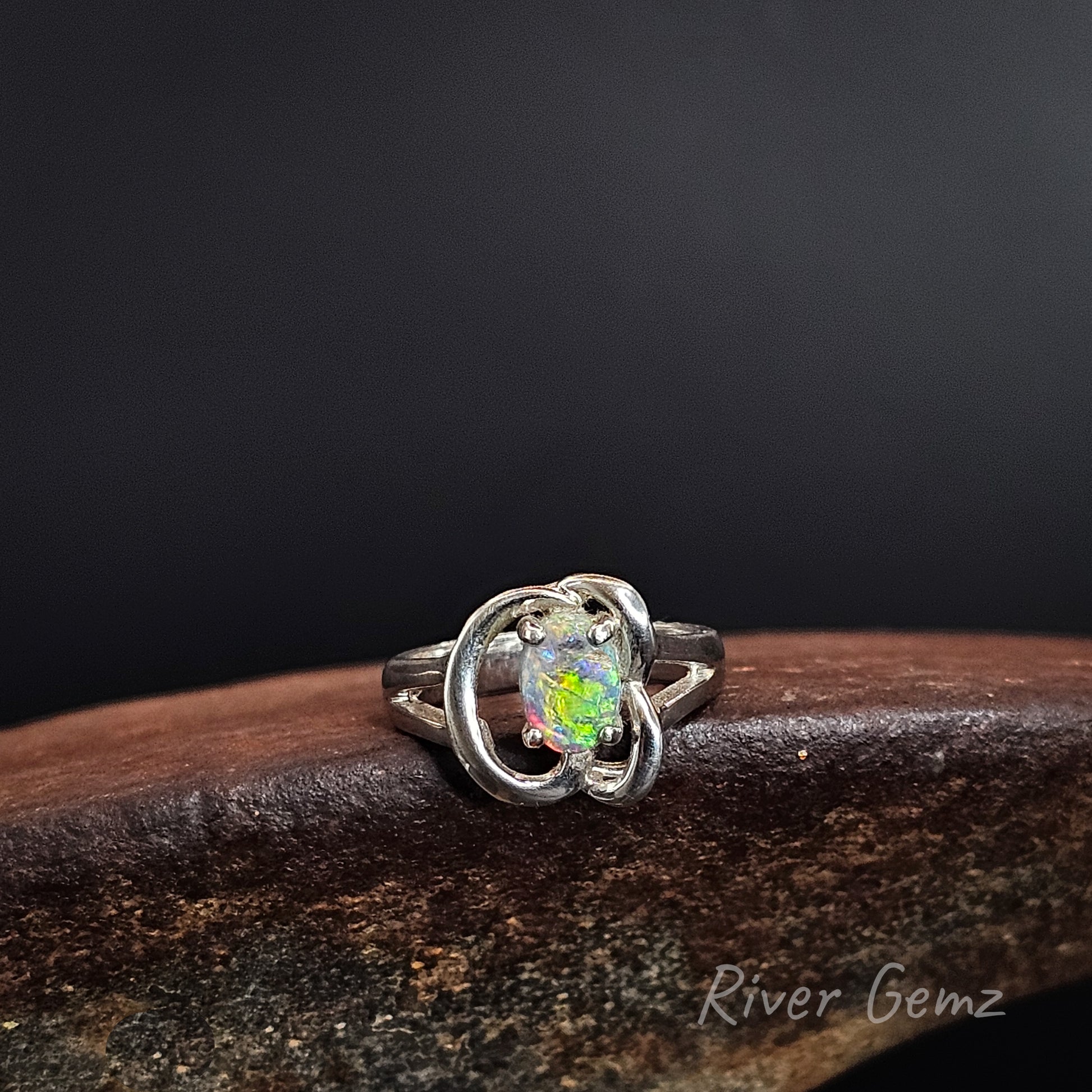 A silver ring featuring a round iridescent opal gemstone in a prong setting, surrounded by a polished silver band with two interlocking curved loops. The ring is displayed on a dark brown wooden surface against a dark gray background, with a 'River Gemz' watermark in the bottom right corner.