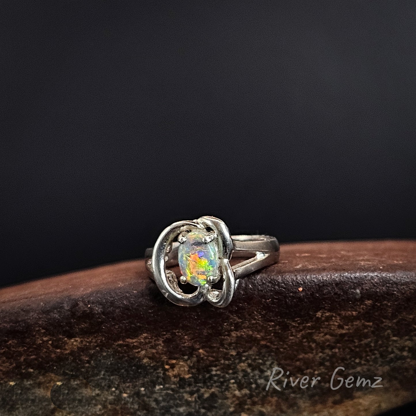 A silver ring featuring an oval cabochon opal with a multicolored play-of-color effect, resting on a dark brown wooden surface against a dark background. The brand name 'River Gemz' is visible as a watermark in the bottom right corner.